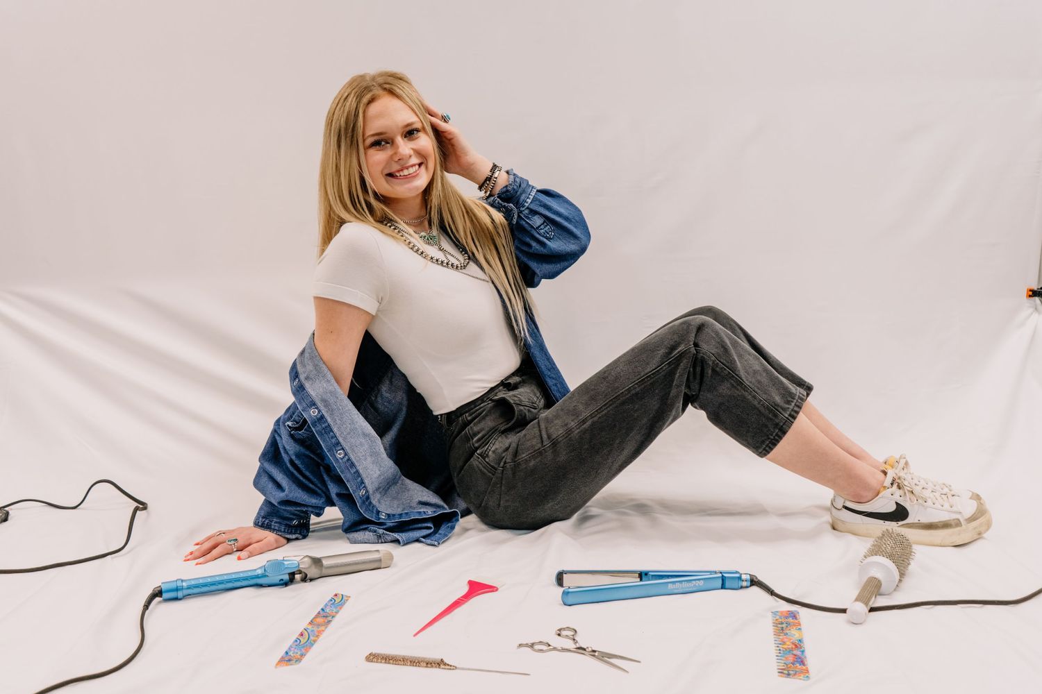 Blonde woman in denim jacket sitting on floor surrounded by hair styling tools including flat irons and scissors.
