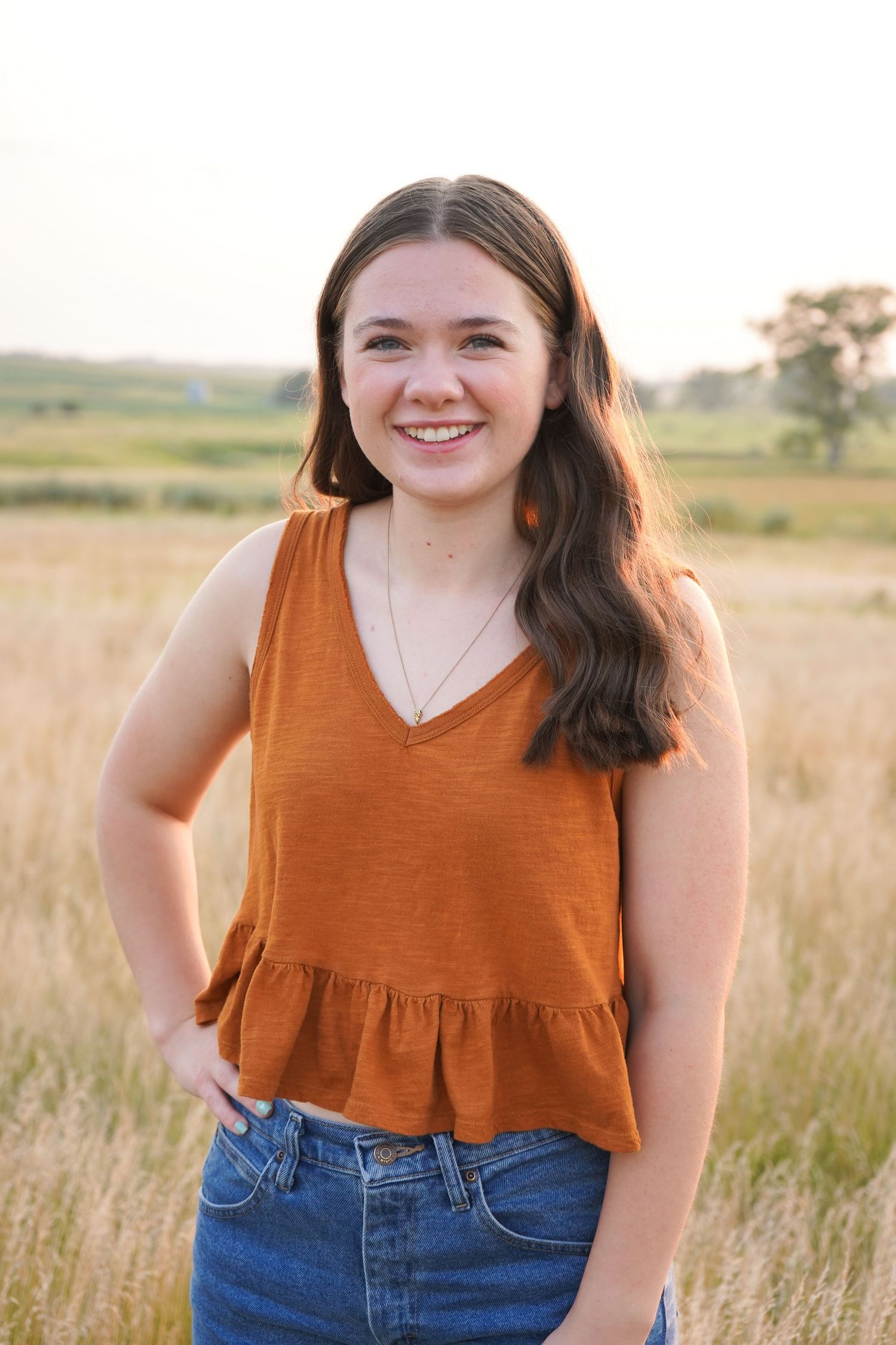 Smiling brunette teen girl in rust orange tank top posing in golden field at sunset.