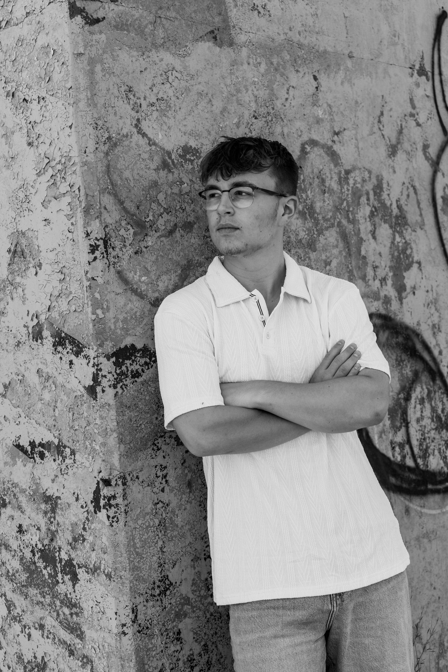 Young man in white polo shirt with arms crossed leaning against graffiti wall, black and white portrait photo.