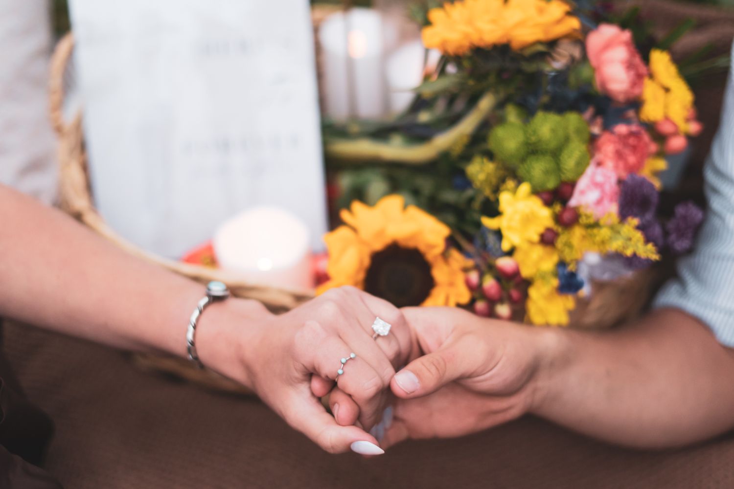 Close-up of couple holding hands displaying a diamond engagement ring with colorful flowers in background.
