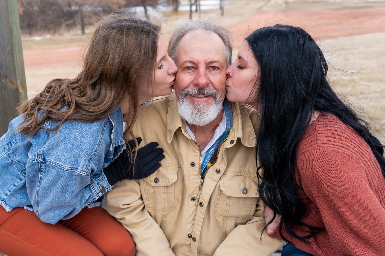 Older bearded man receiving kisses on cheeks from two younger women outdoors in winter.