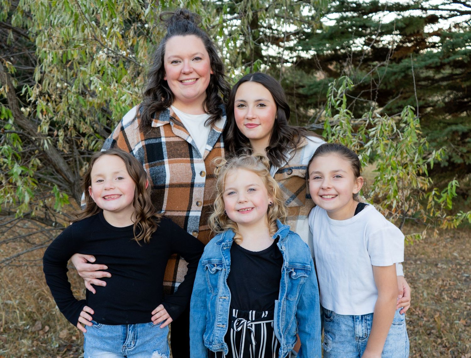 Mother posing outdoors with four daughters of varying ages surrounded by green trees.