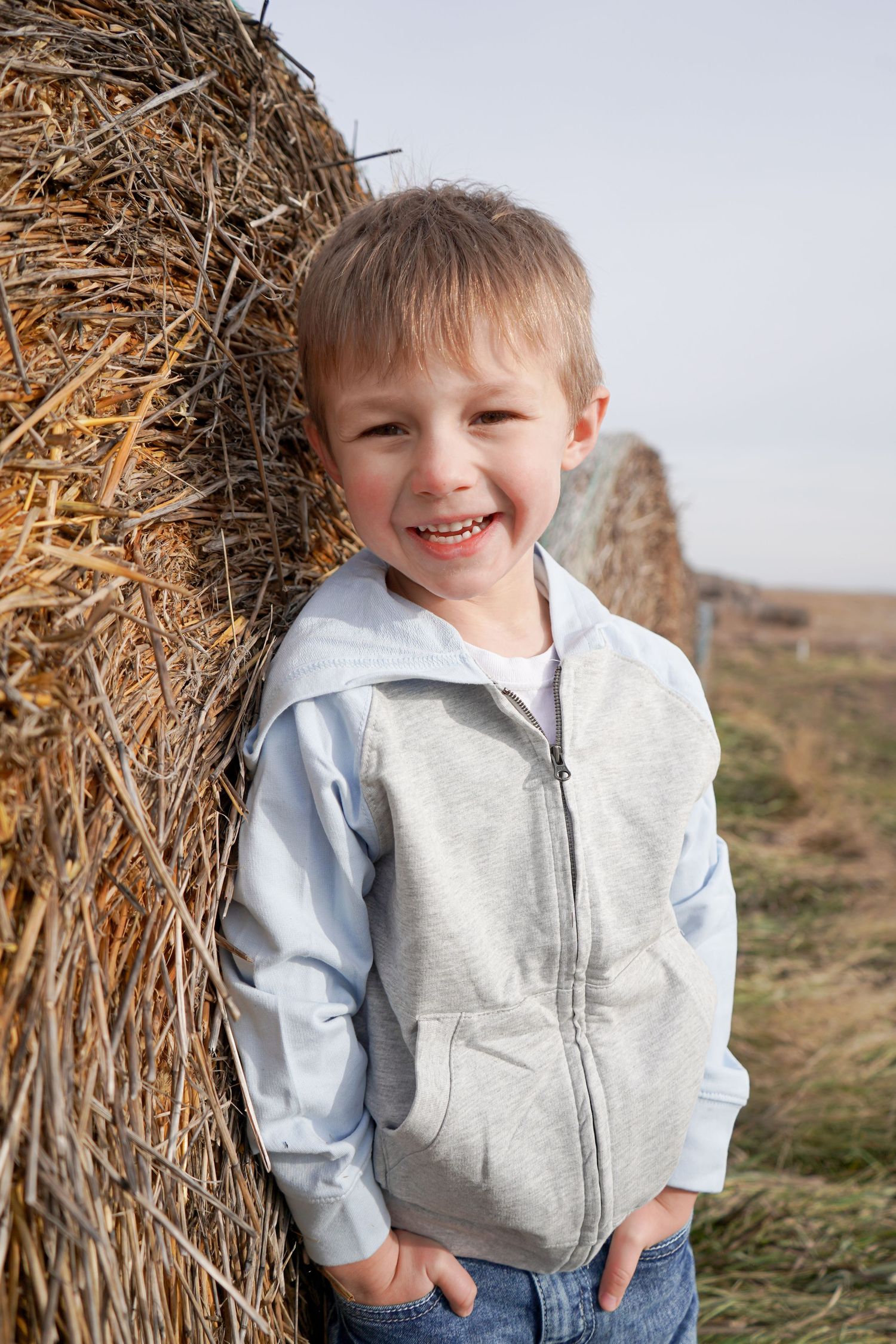 Cheerful young boy in gray hoodie leans against a large hay bale in an open field.