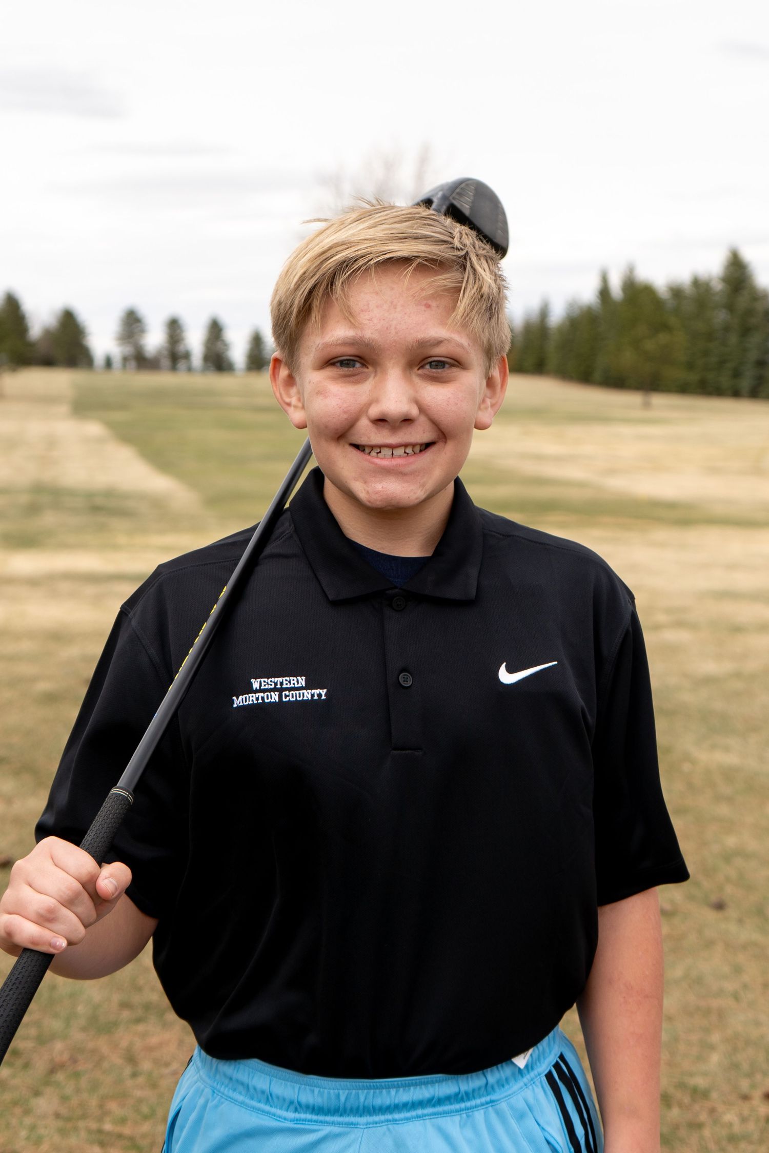 A smiling young boy in a black Nike polo holds a golf club on a golf course wearing blue shorts.