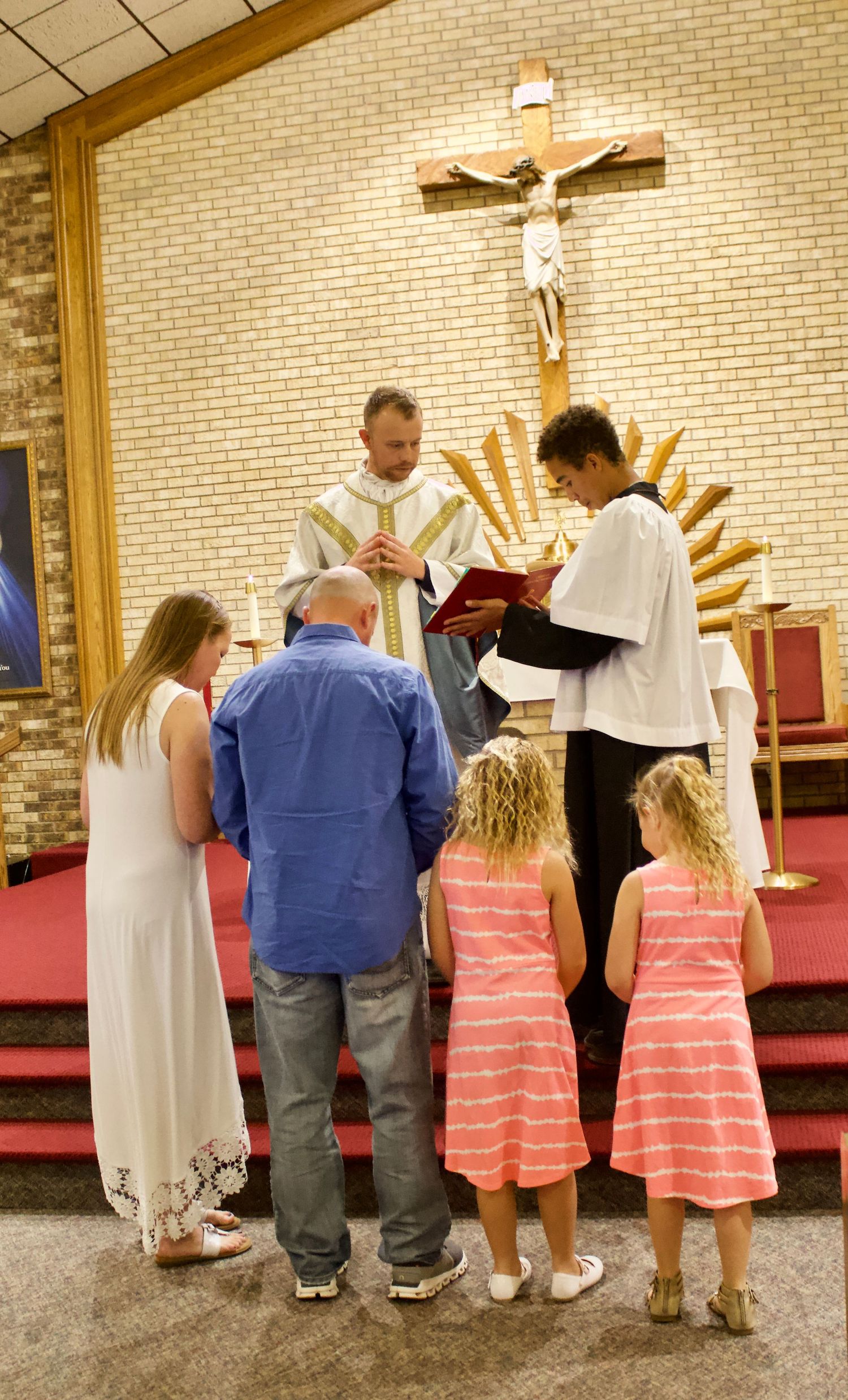 Family gathered at church altar with priest and altar server during a Catholic religious ceremony.