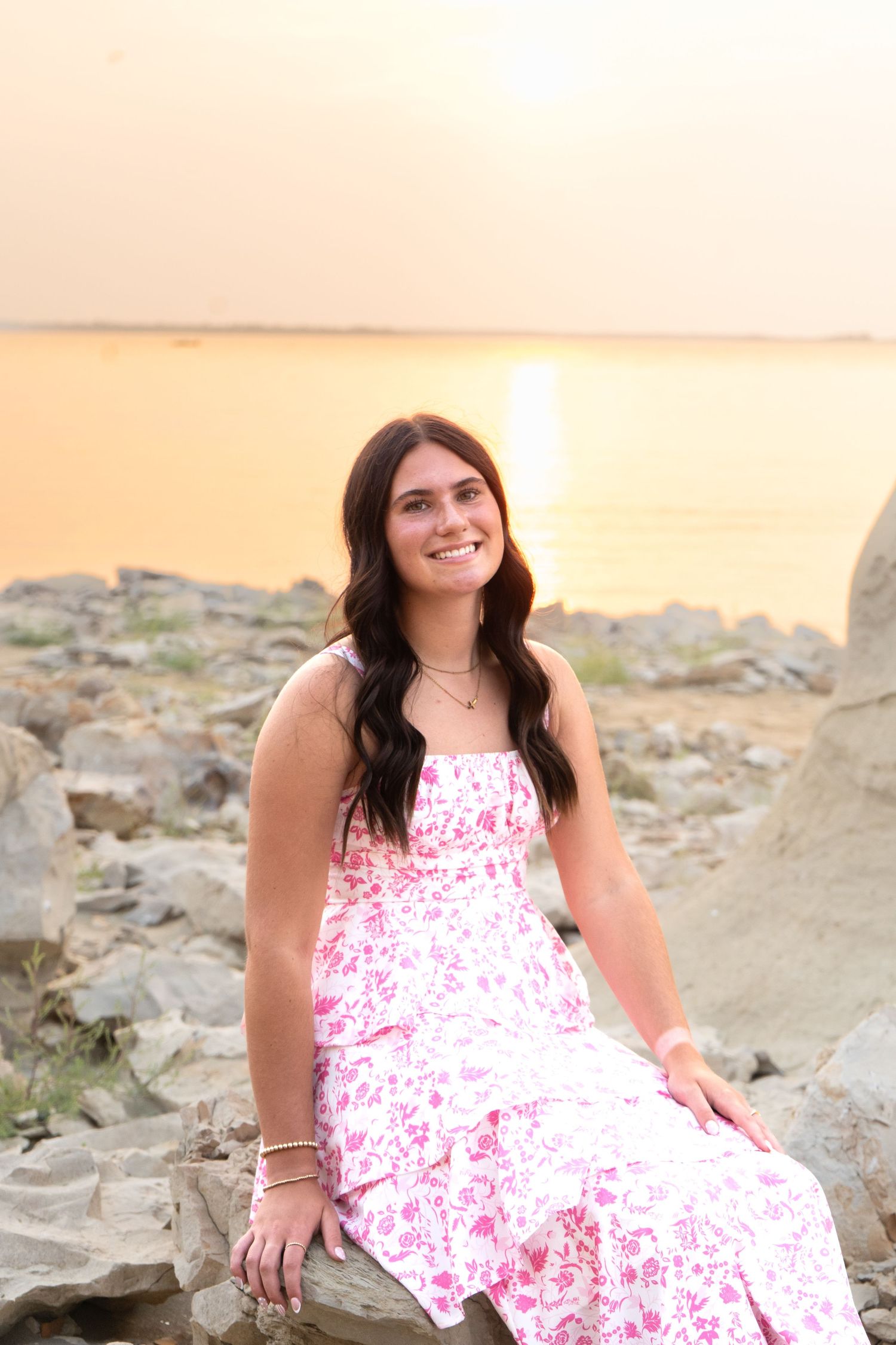 Smiling brunette in pink floral sundress sits on rocks by lake during golden sunset.