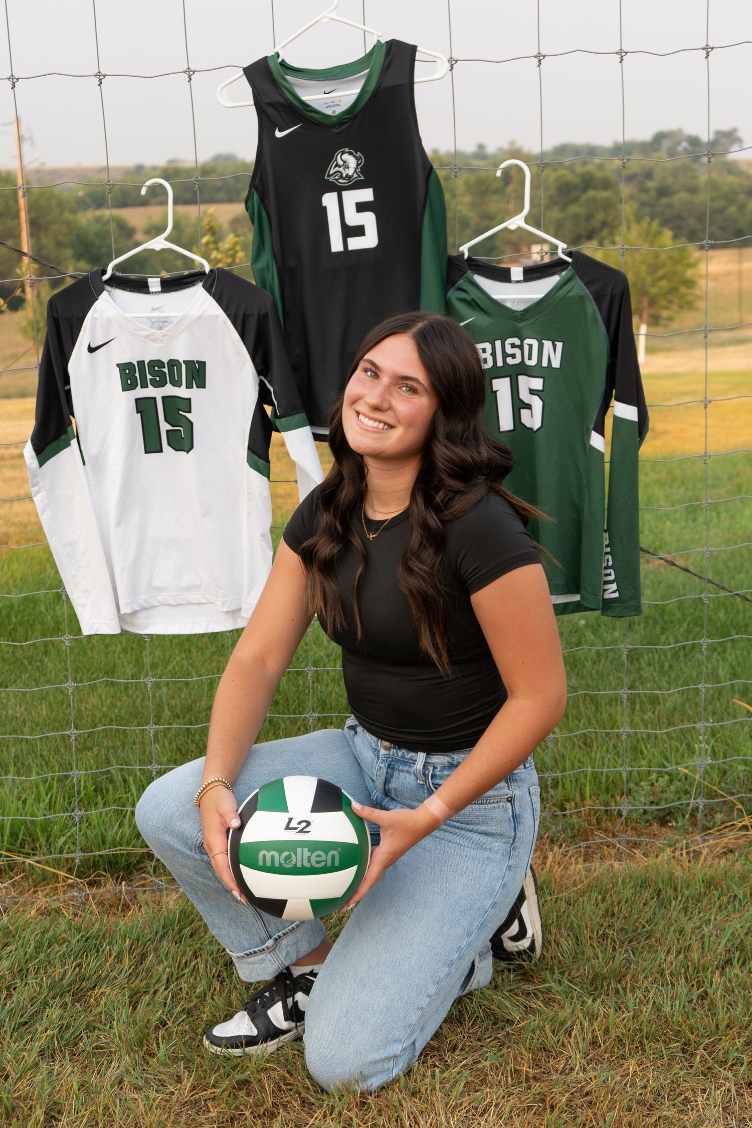 Smiling female athlete holds Molten volleyball in front of three Bison number 15 jerseys outdoors.