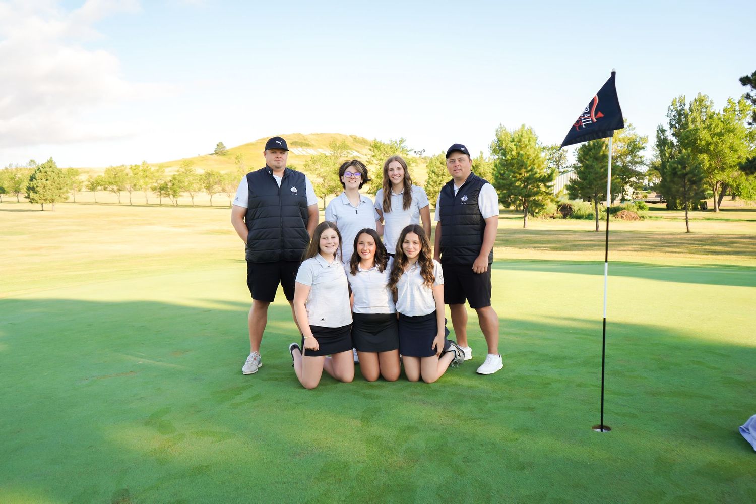 A high school golf team poses together on a putting green next to a flag on a sunny day.