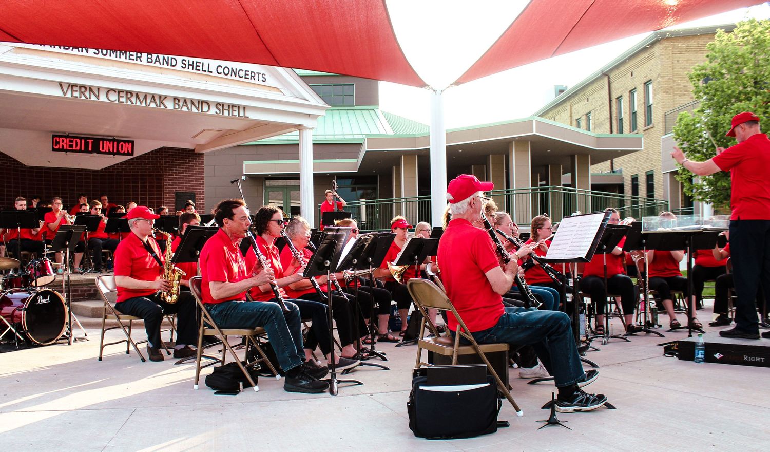 Community band in red uniforms performing at the Vern Cernak Band Shell outdoor concert.