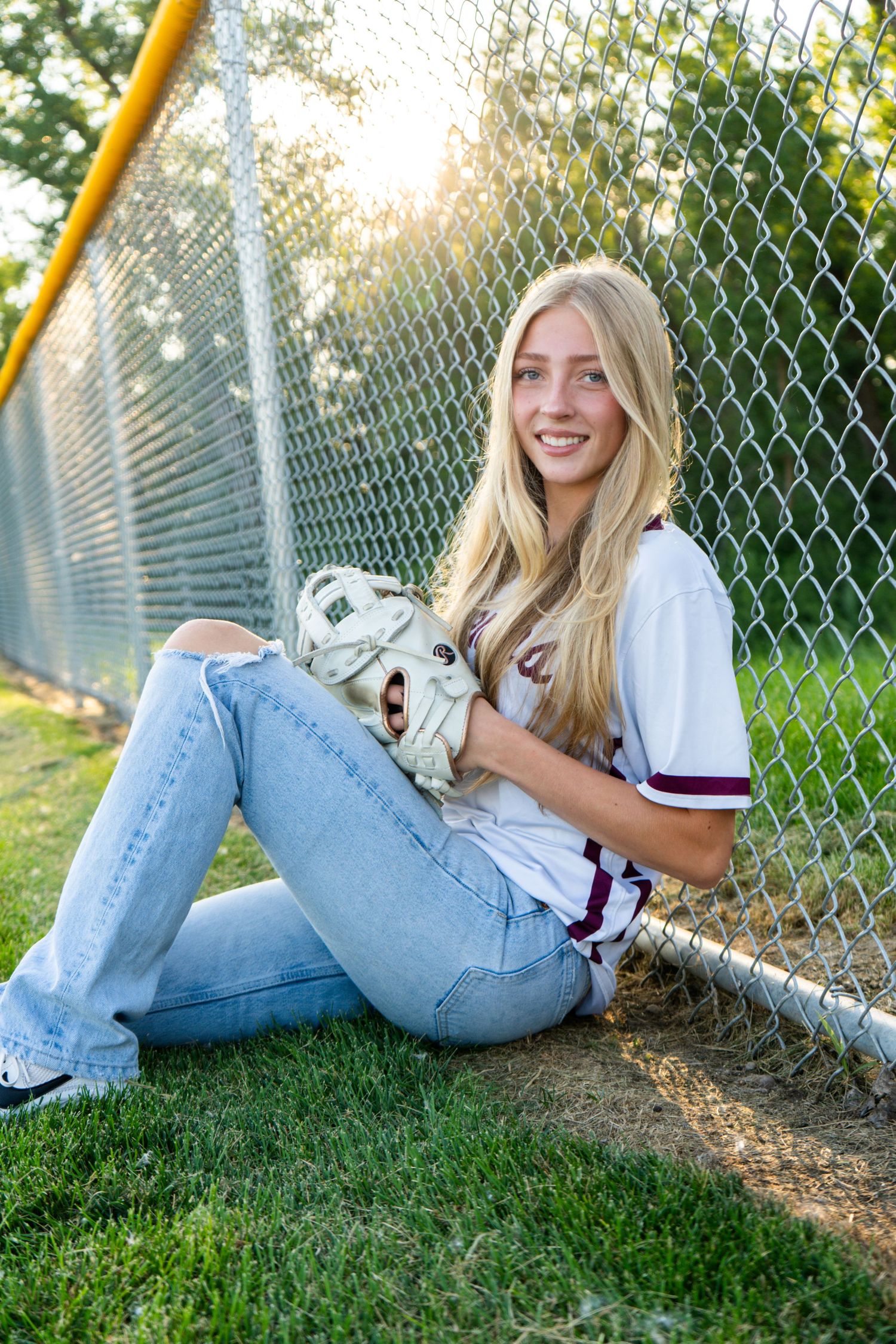 Smiling blonde softball player sitting against chain-link fence holding a catcher glove in golden light.