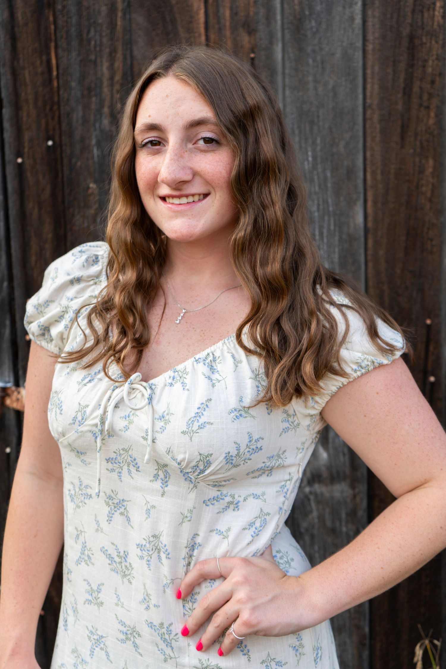 Smiling young woman with wavy brown hair wearing a floral white dress standing in front of a wooden barn wall.