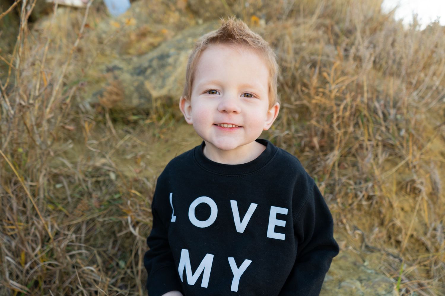 Smiling toddler boy in black Love My sweatshirt standing in natural outdoor setting.