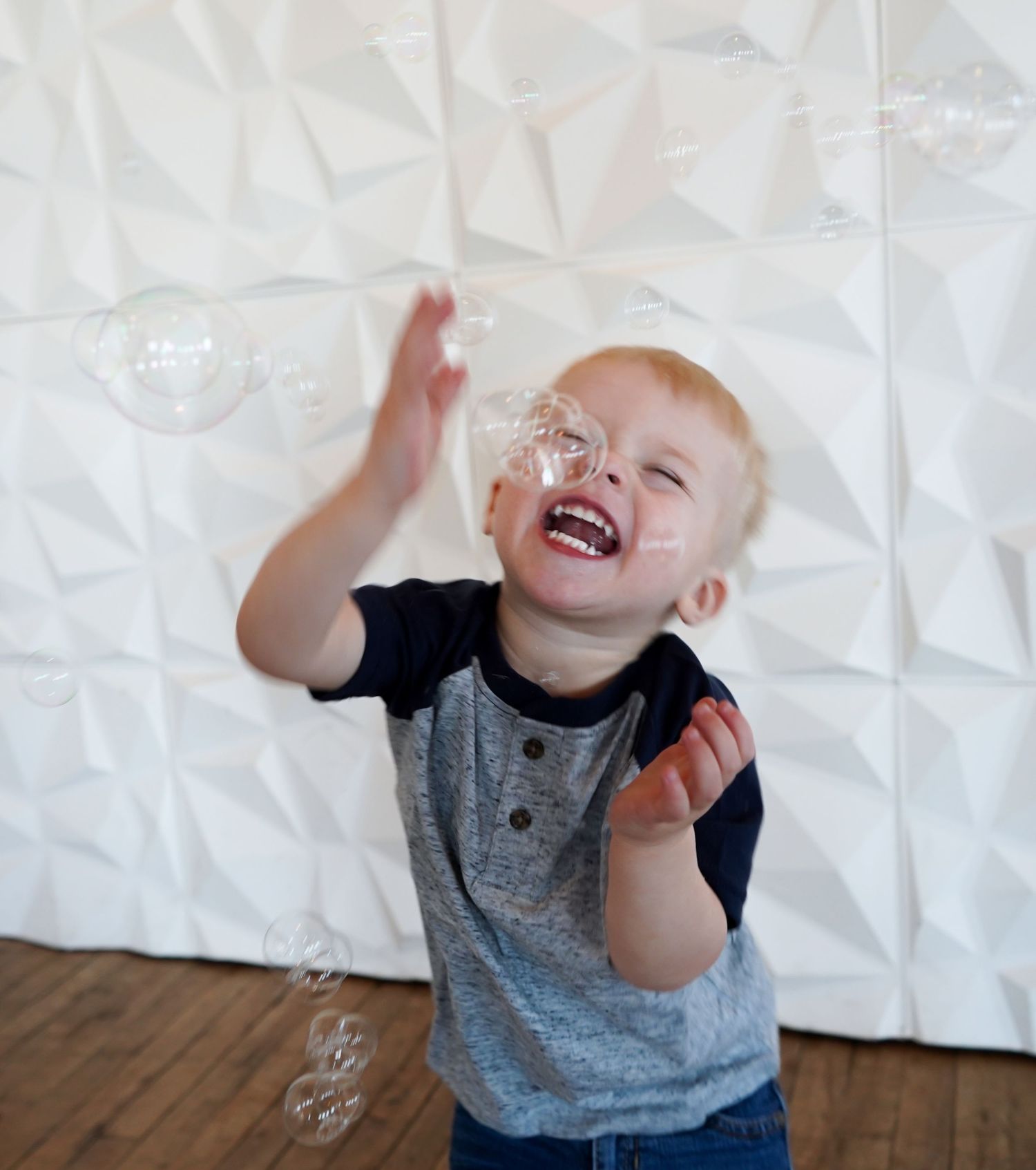 Joyful toddler boy laughing and reaching up to catch soap bubbles against a white textured wall.