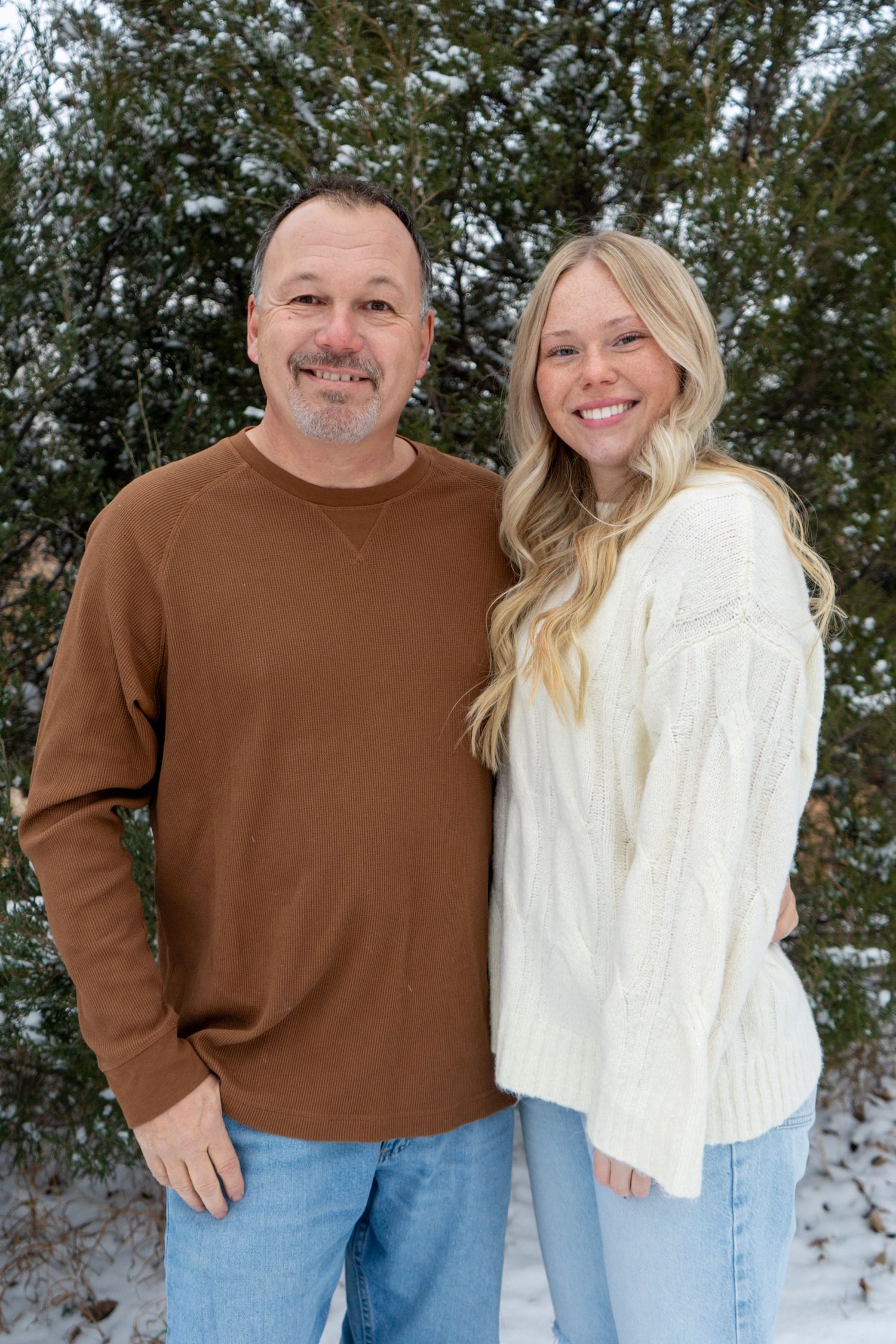 Man and young blonde woman smiling together outdoors in winter with snow-covered evergreen trees.