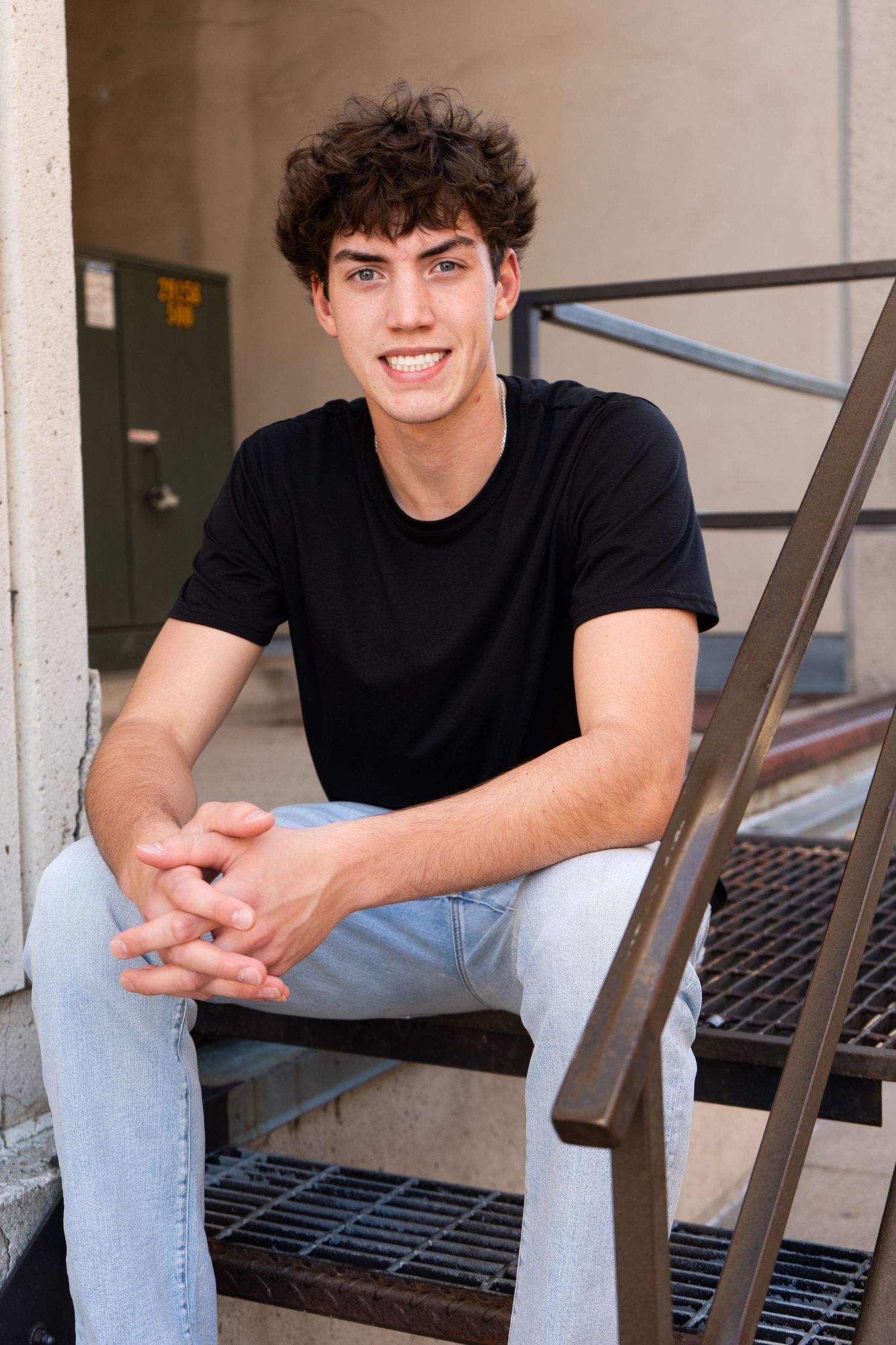 Smiling teenage boy in black t-shirt and jeans sitting on metal stairs outdoors.