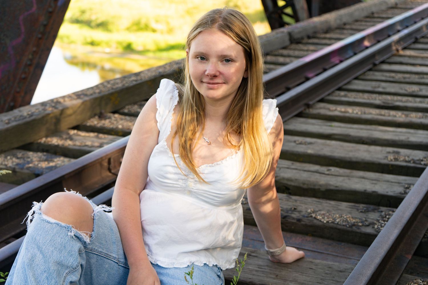Blonde teen girl in white top and ripped jeans sits on rustic railroad bridge.