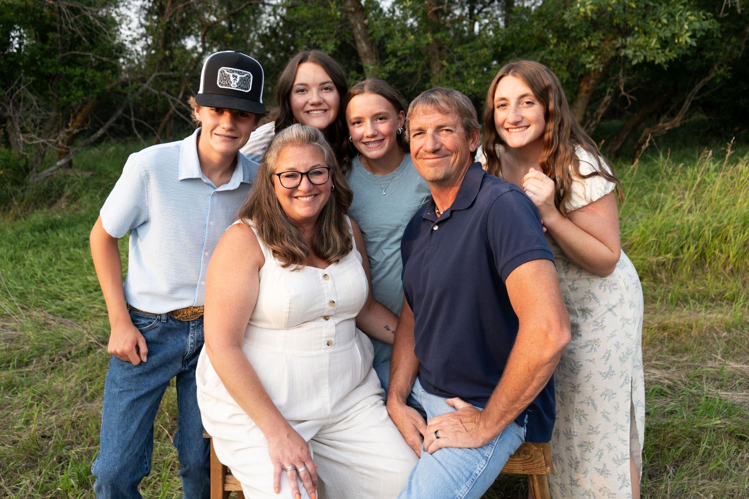 A family of six poses together outdoors on a wooden bench surrounded by lush greenery.