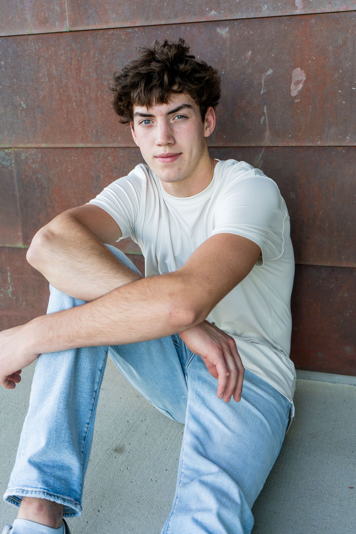 Teen boy in white t-shirt and jeans sitting against rustic metal wall outdoors.