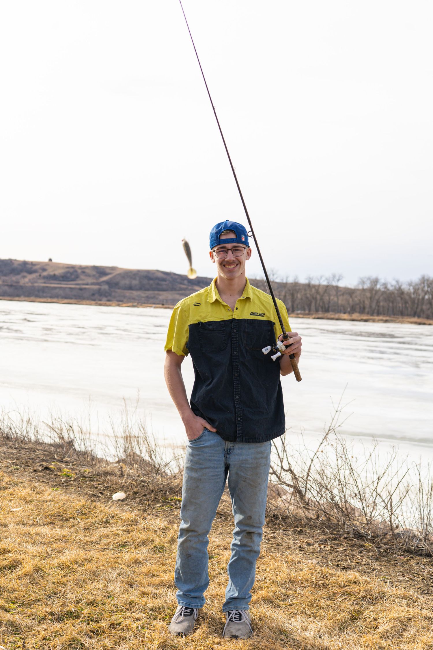 Smiling young man holding fishing rod near frozen river on overcast spring day.