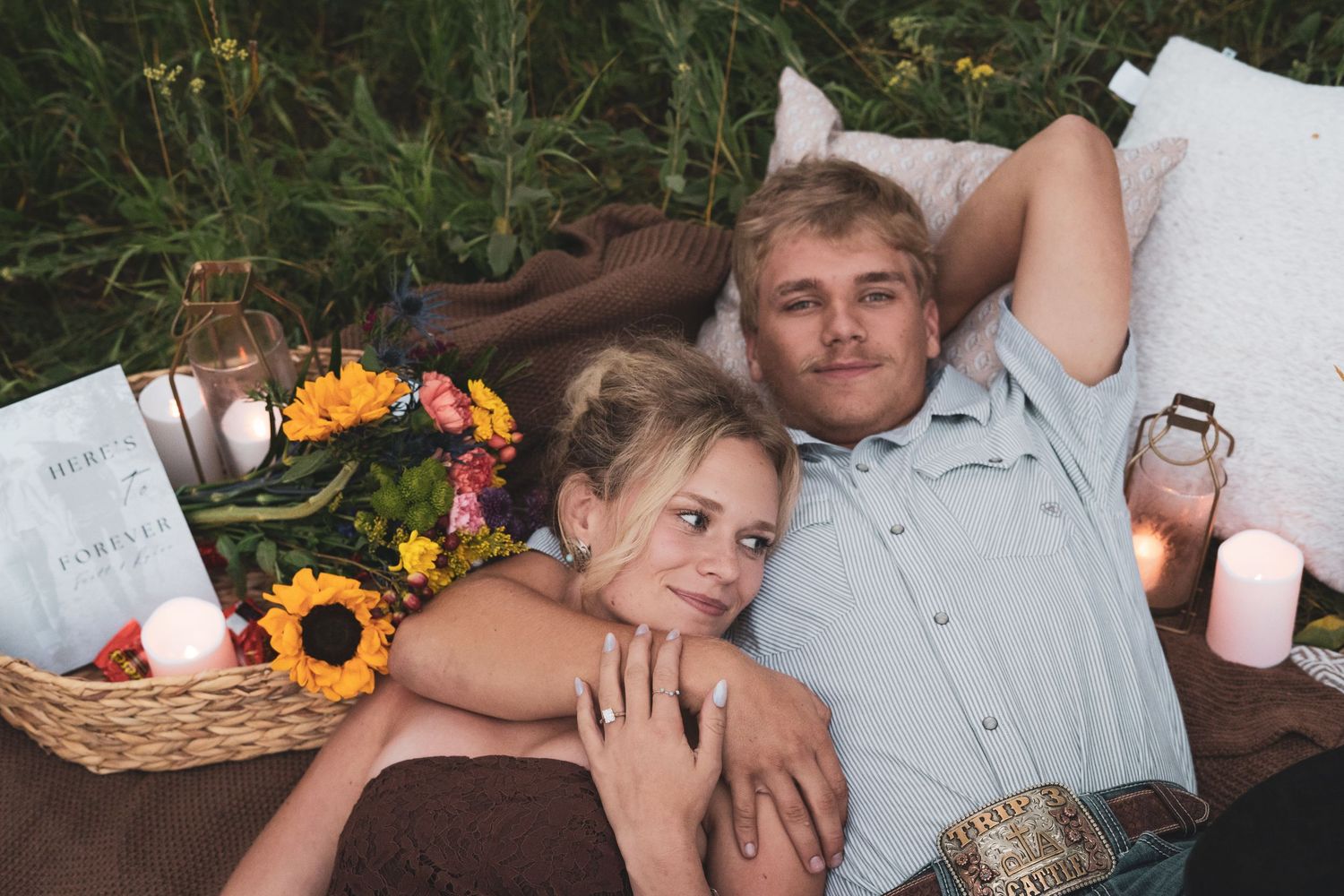 Couple lying in grass field on picnic blanket surrounded by sunflowers, candles, and a wicker basket.