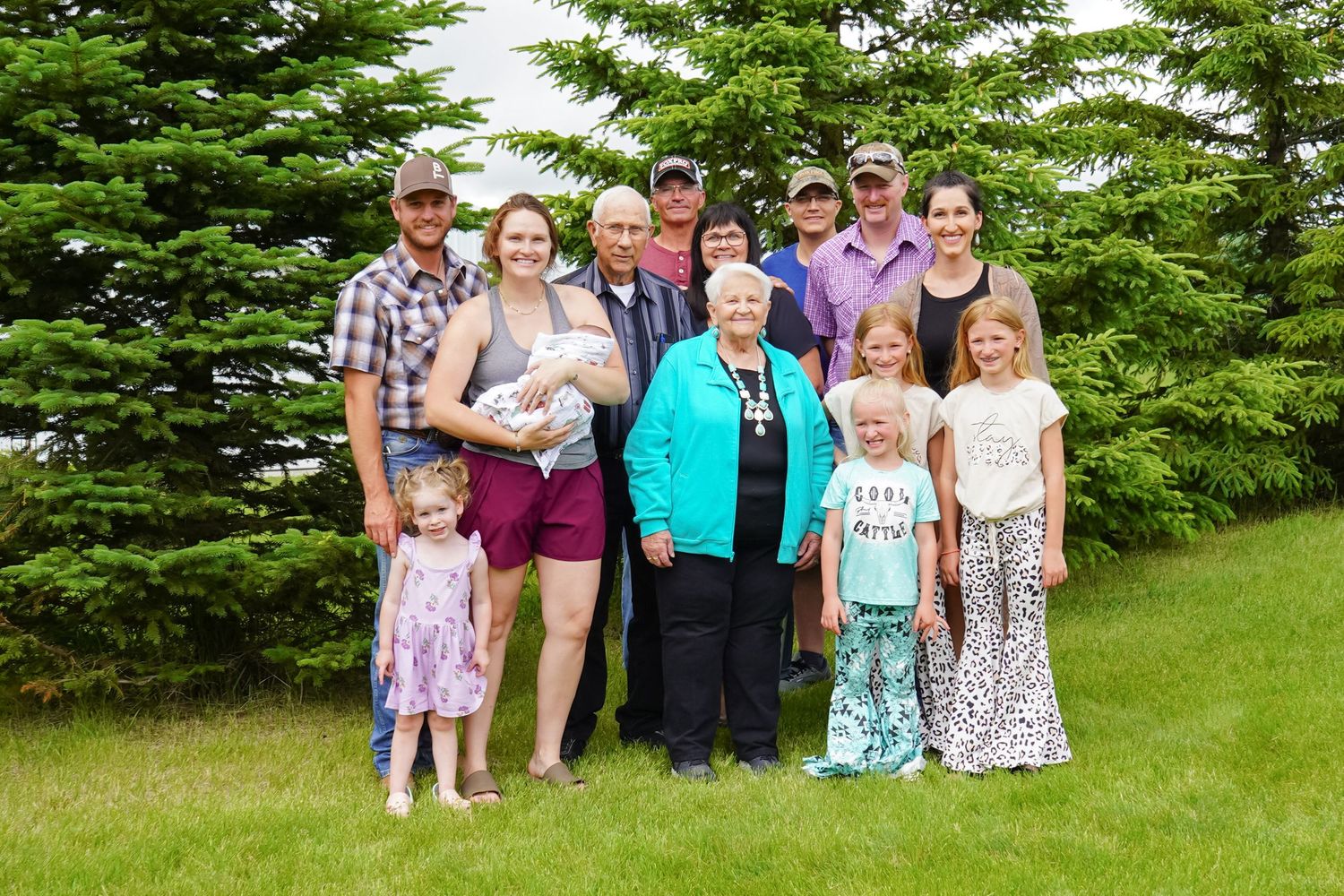 Multi-generational family group smiling together on a green lawn with tall evergreen trees behind them.