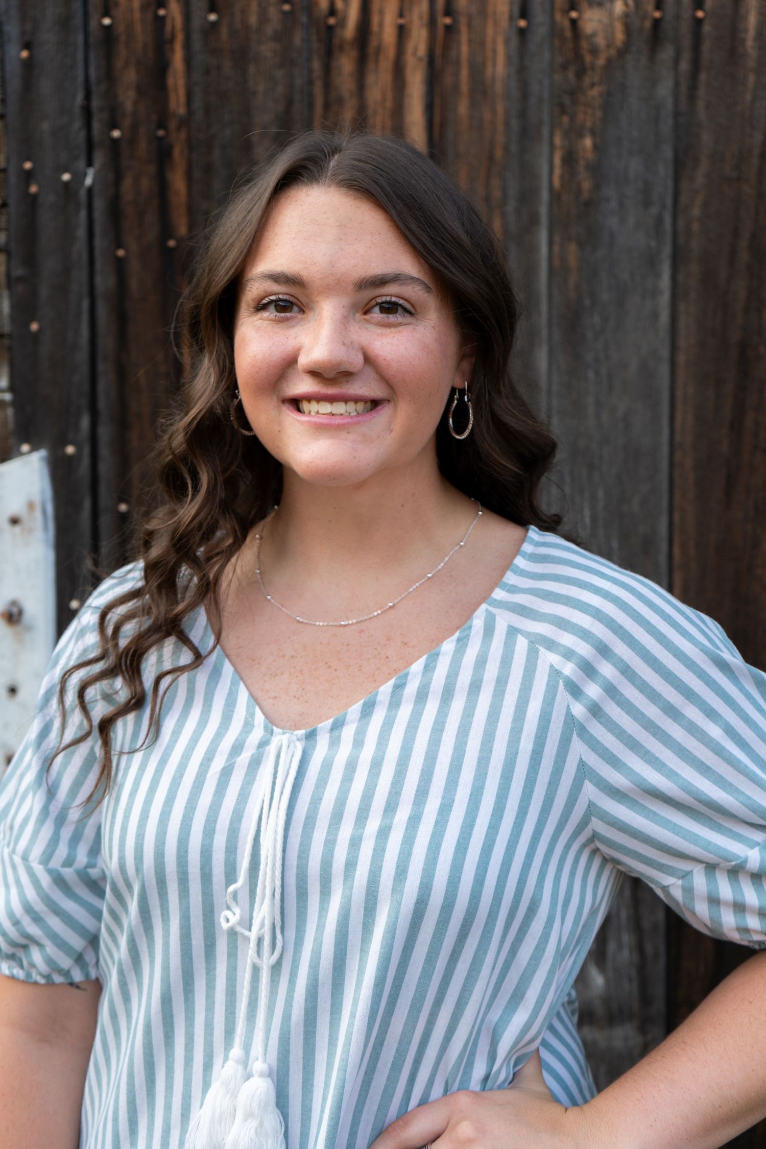 Smiling young woman with wavy brown hair wearing a blue and white striped blouse standing by a rustic wooden wall.