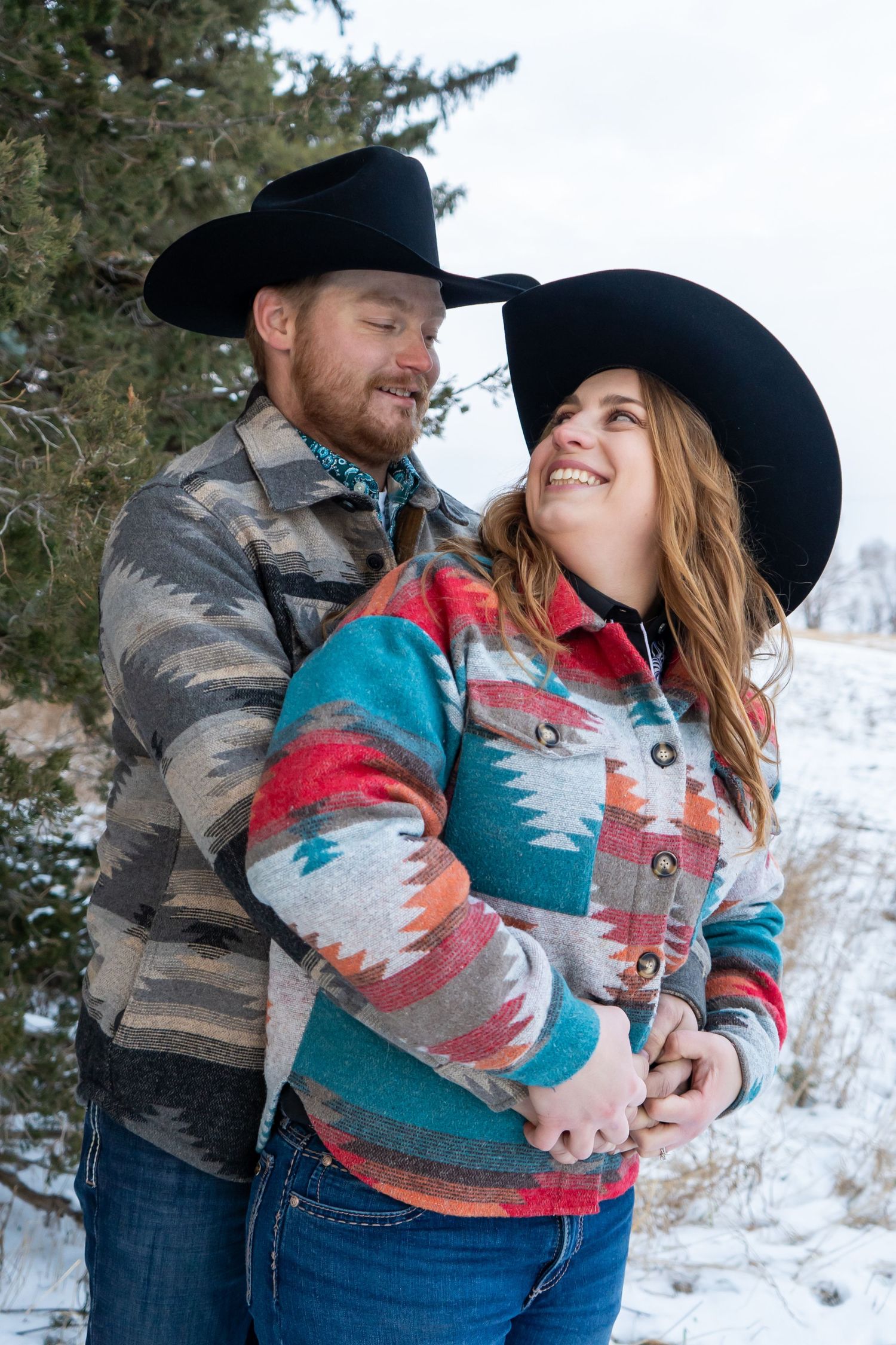 Happy couple in western Aztec print jackets and black cowboy hats embracing in a winter snow scene.