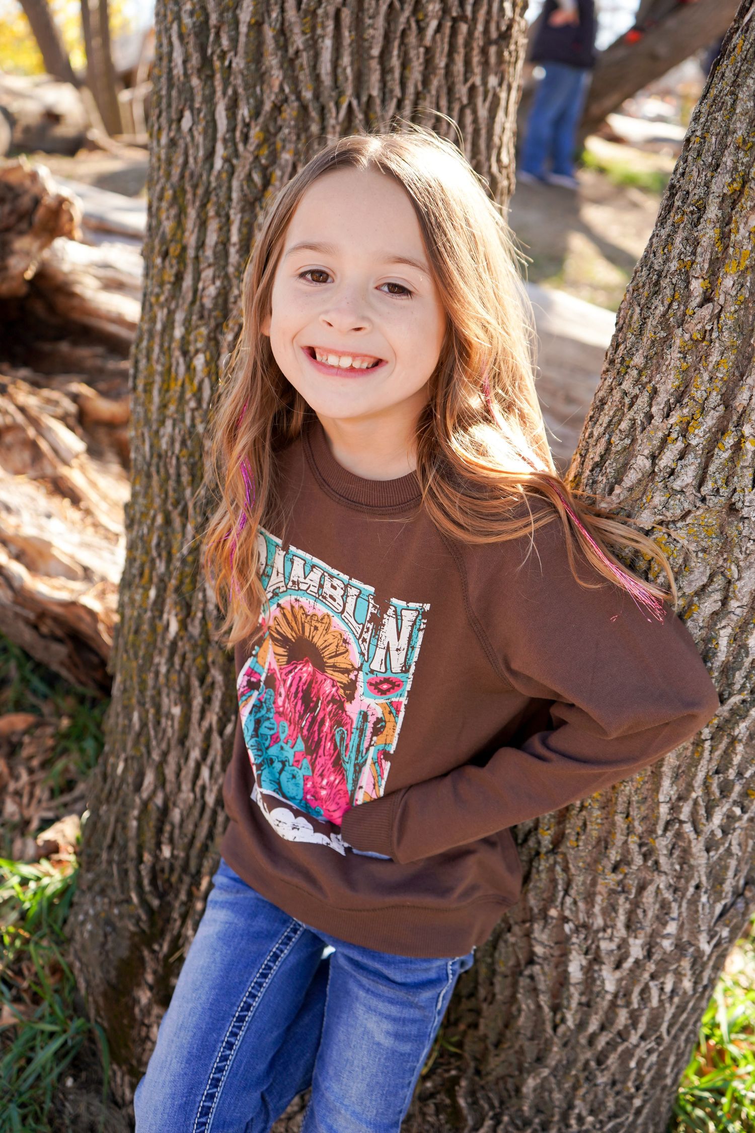 Young girl smiling in brown Ramblin graphic sweatshirt leaning against tree trunk outdoors.
