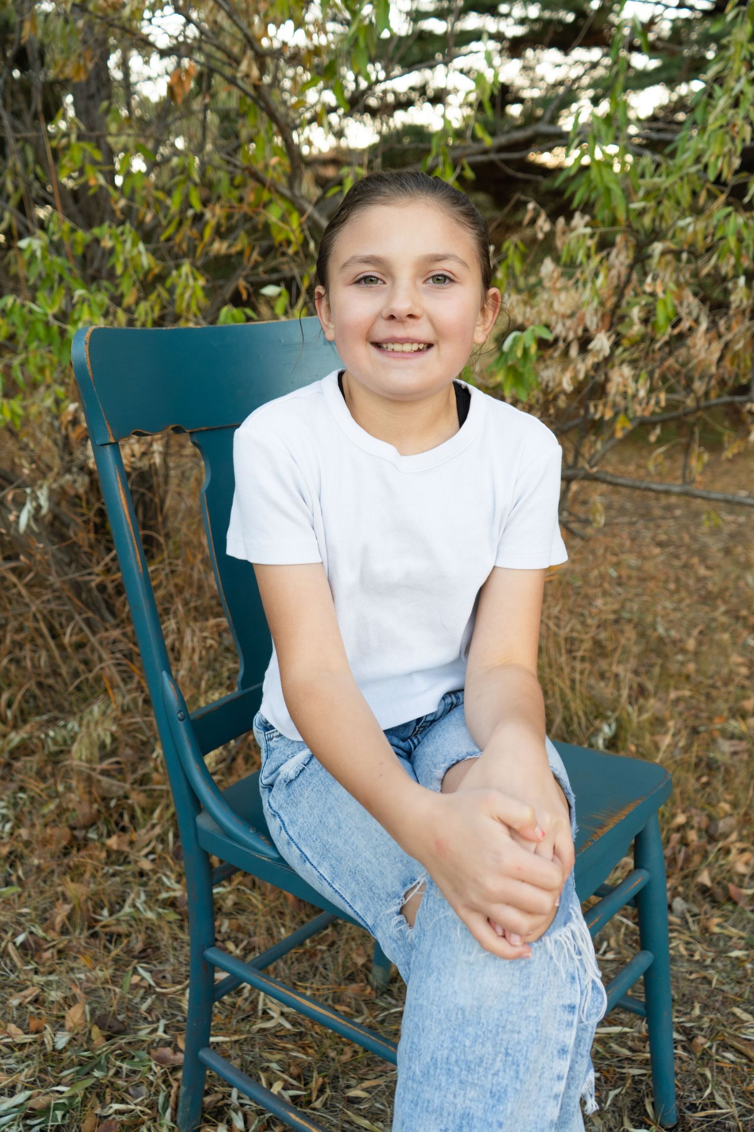 Smiling preteen girl in white t-shirt and jeans sitting on teal chair outdoors.