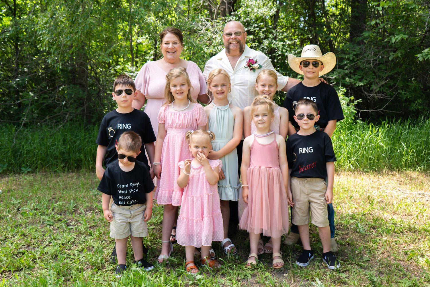 A couple poses with a group of children wearing sunglasses and ring bearer shirts at an outdoor wedding.