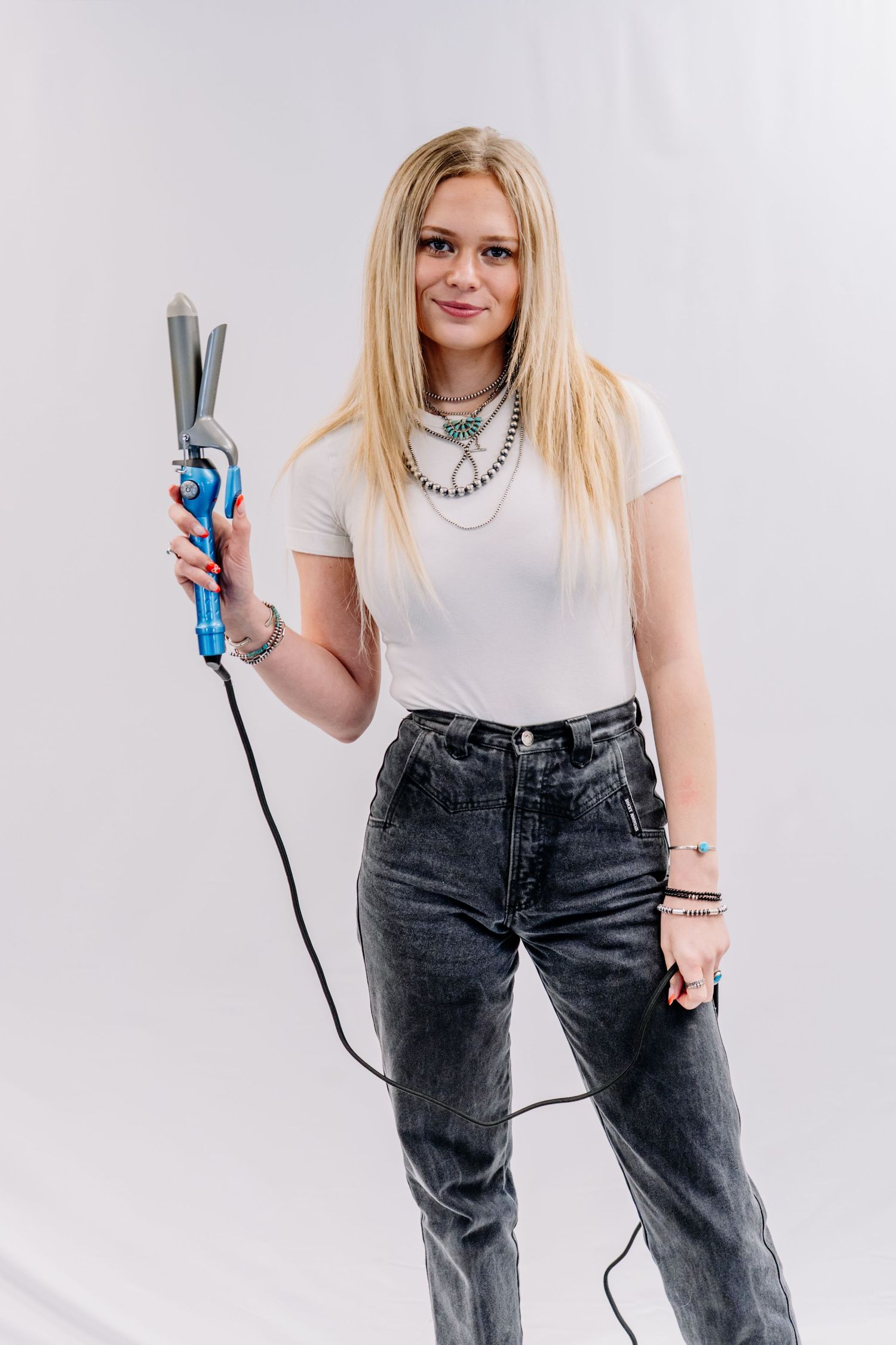 Blonde female hairstylist posing with blue curling iron in white studio setting.
