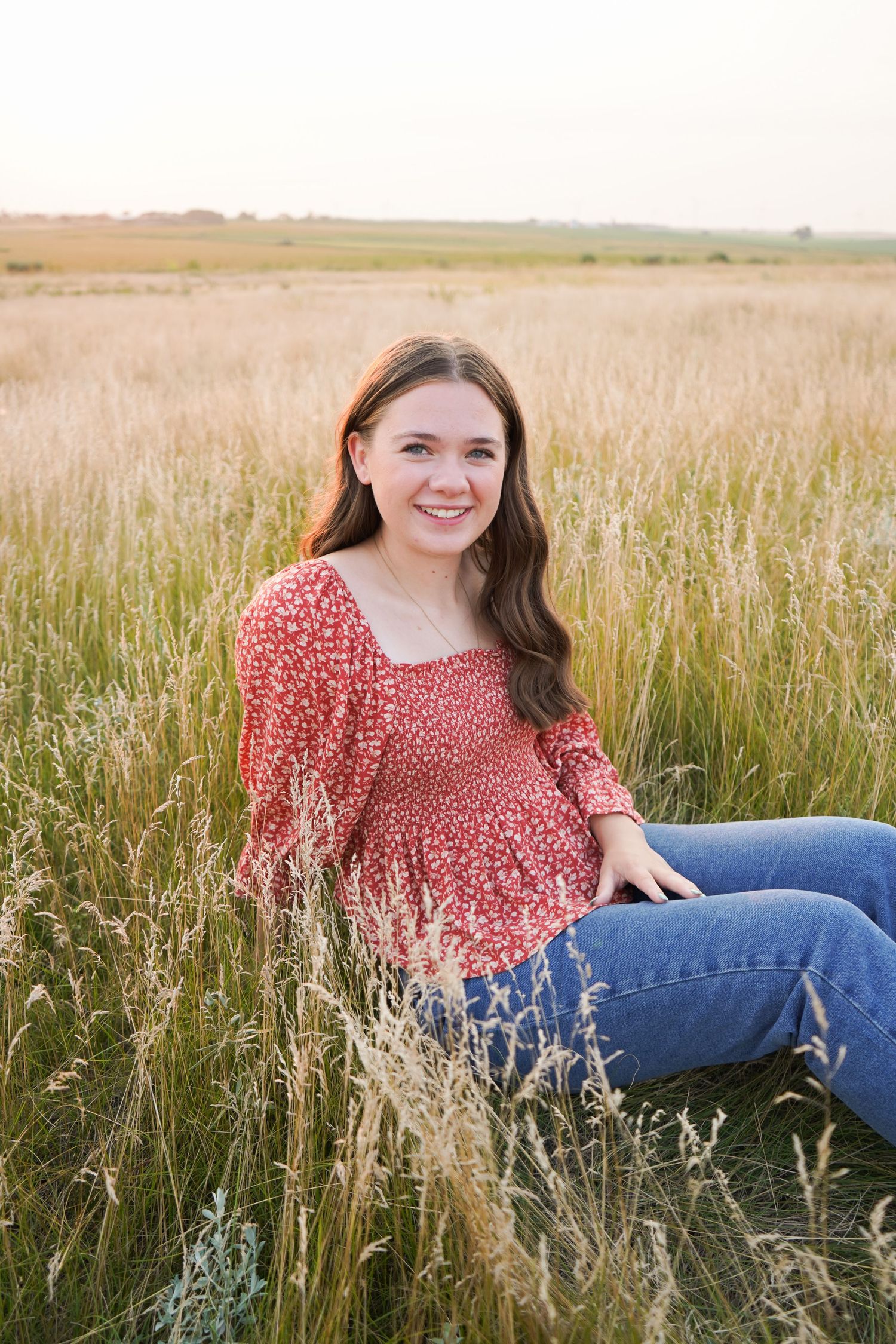 Young woman in red floral top and jeans sitting in tall grass field at golden hour.