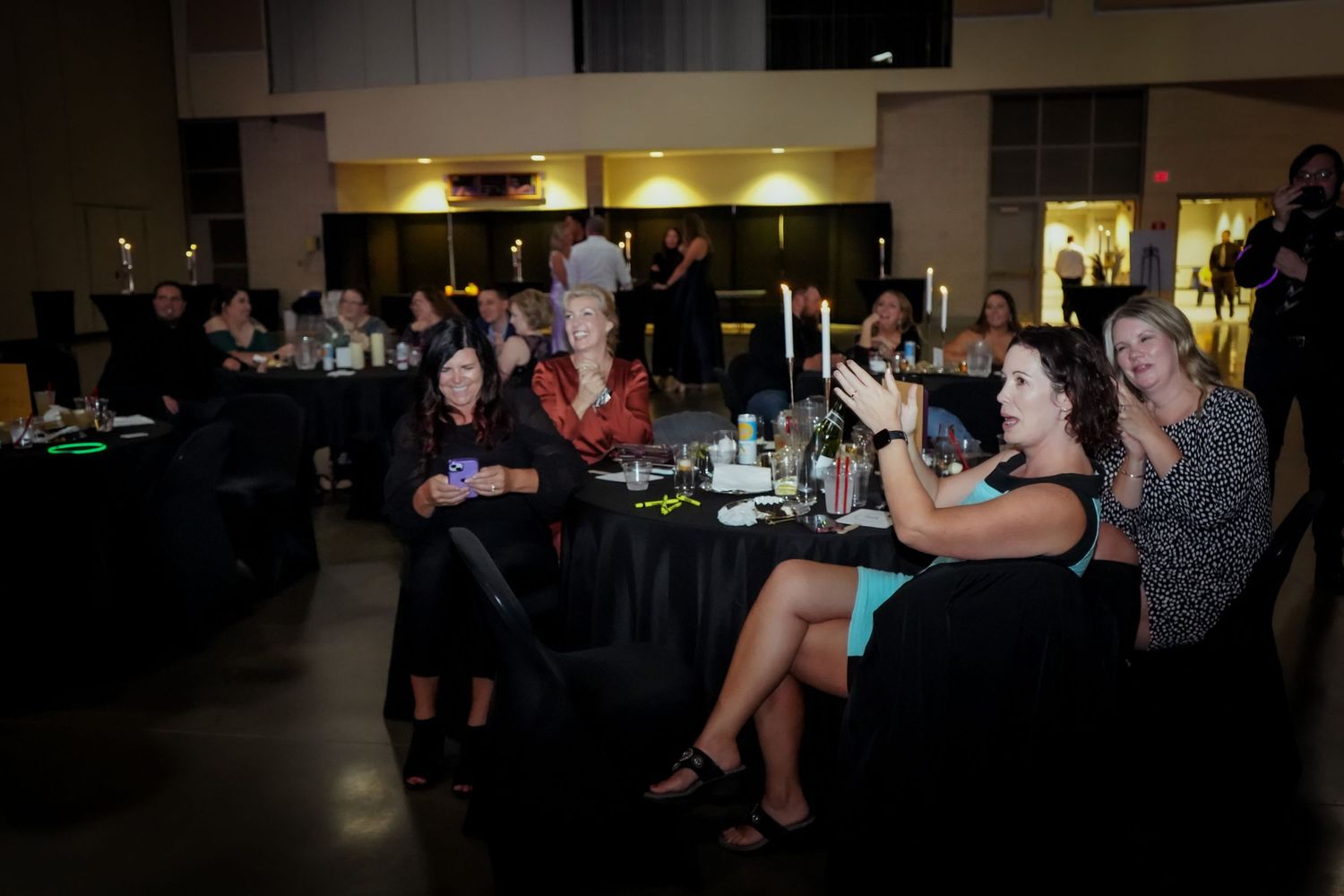 Guests seated at black-draped tables applauding at a dimly lit formal evening event or gala.