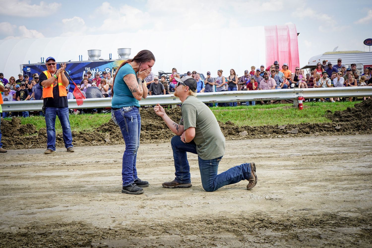Man kneels proposing to woman on dirt track at crowded outdoor event.
