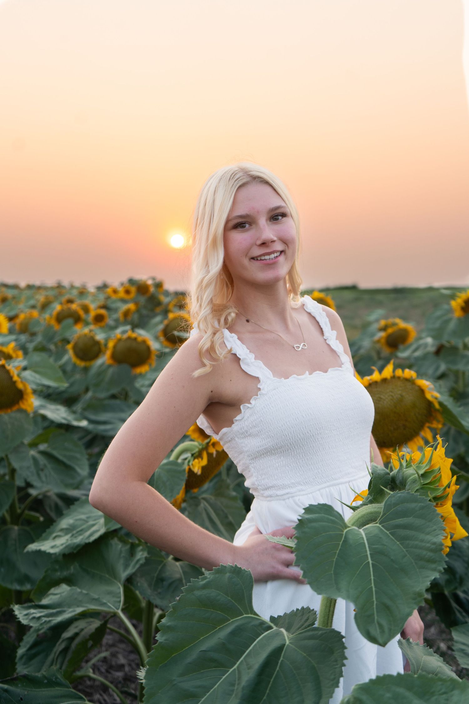 Smiling blonde woman in white sundress posing in sunflower field at vibrant sunset.
