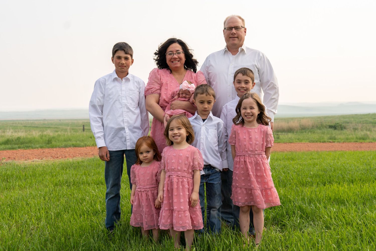 Large family of eight posing outdoors in a green field, parents with six children dressed in pink and white.