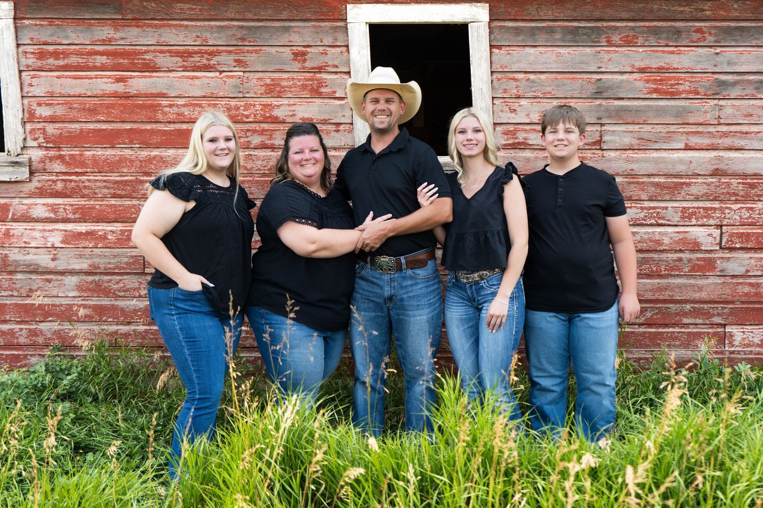 Five family members in black tops and jeans smile together in front of a rustic red barn.