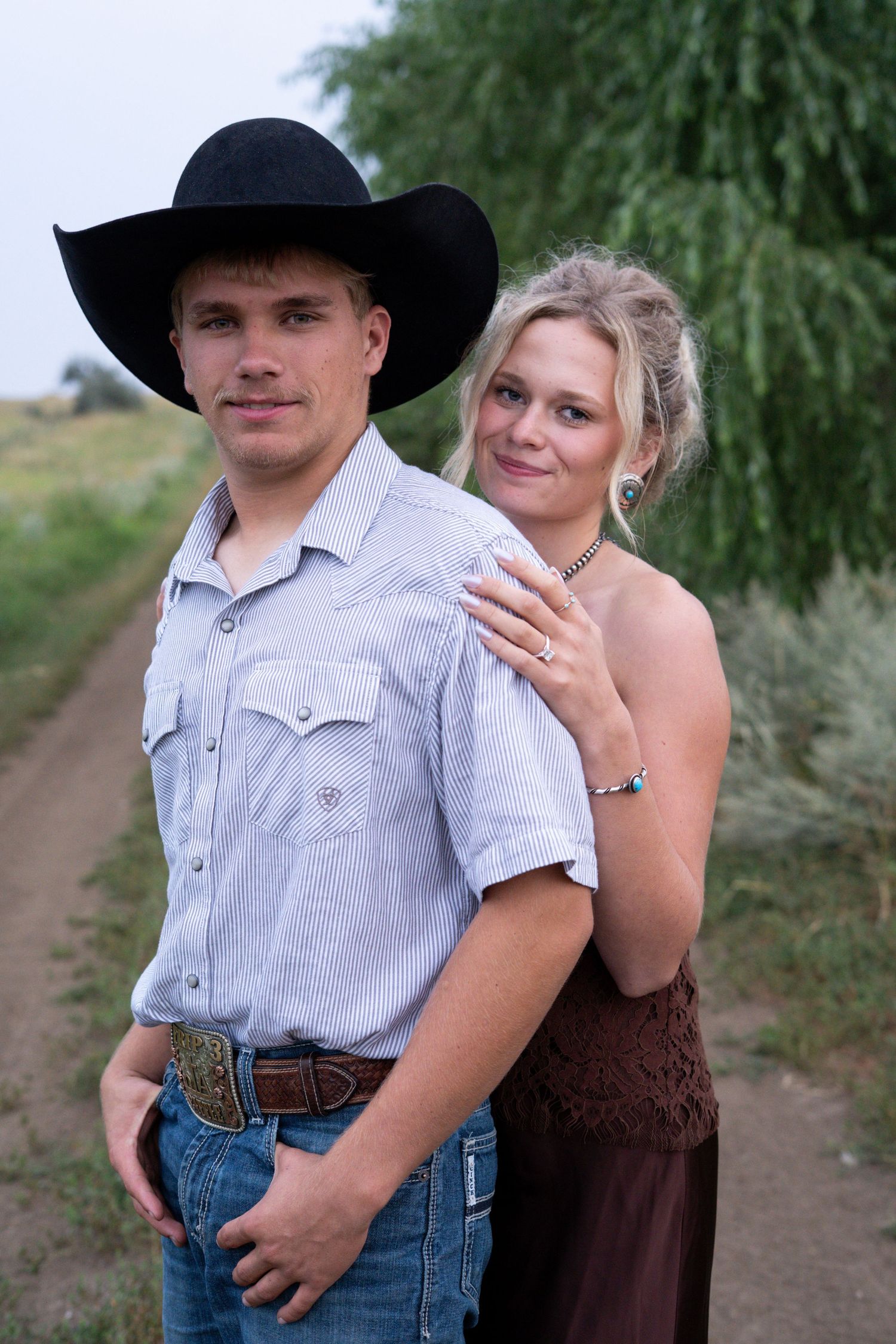 Young couple in western attire posing on a rural dirt road, man wearing black cowboy hat and woman in brown dress.