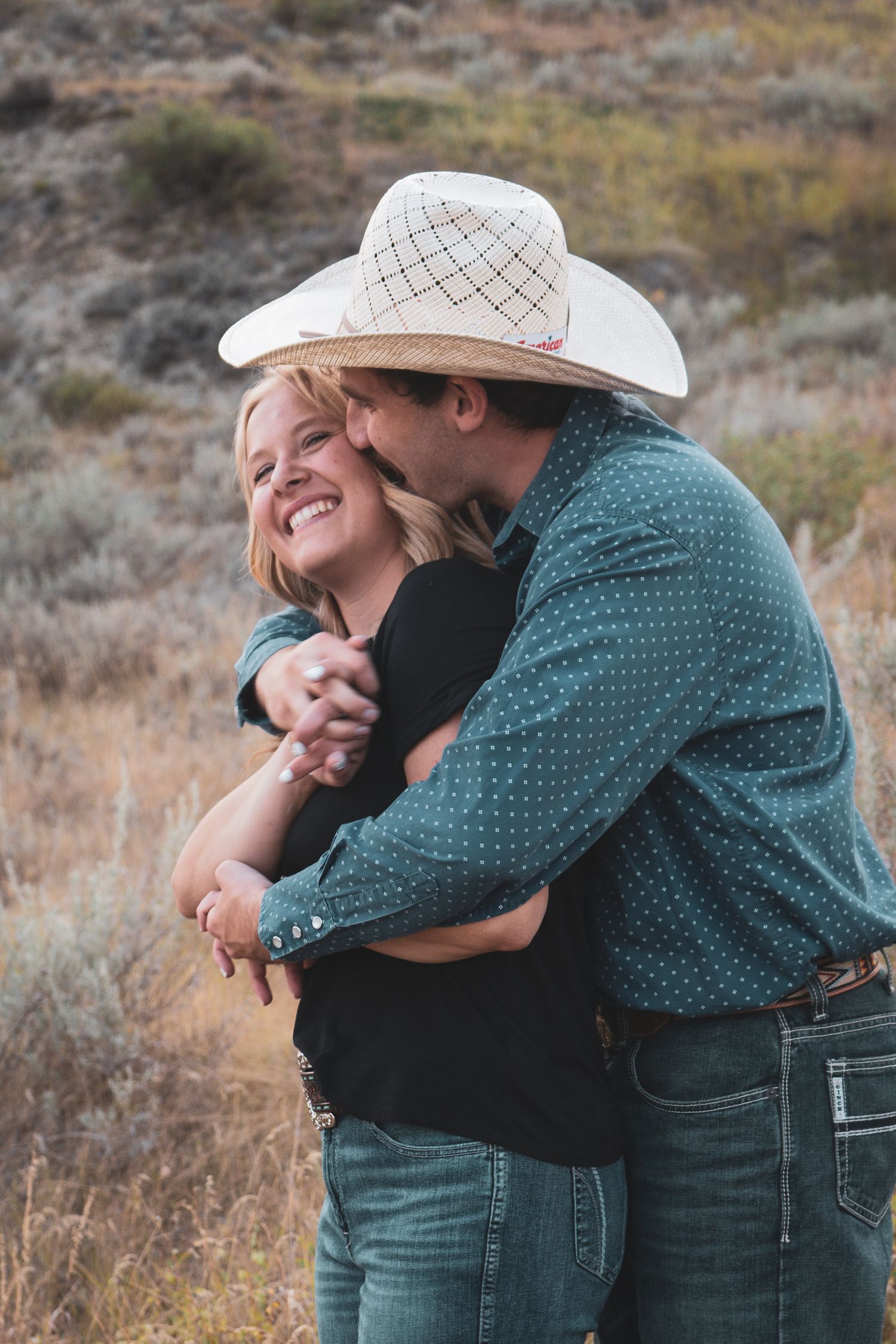 Laughing blonde woman being embraced and kissed by man in white cowboy hat in a sagebrush landscape.