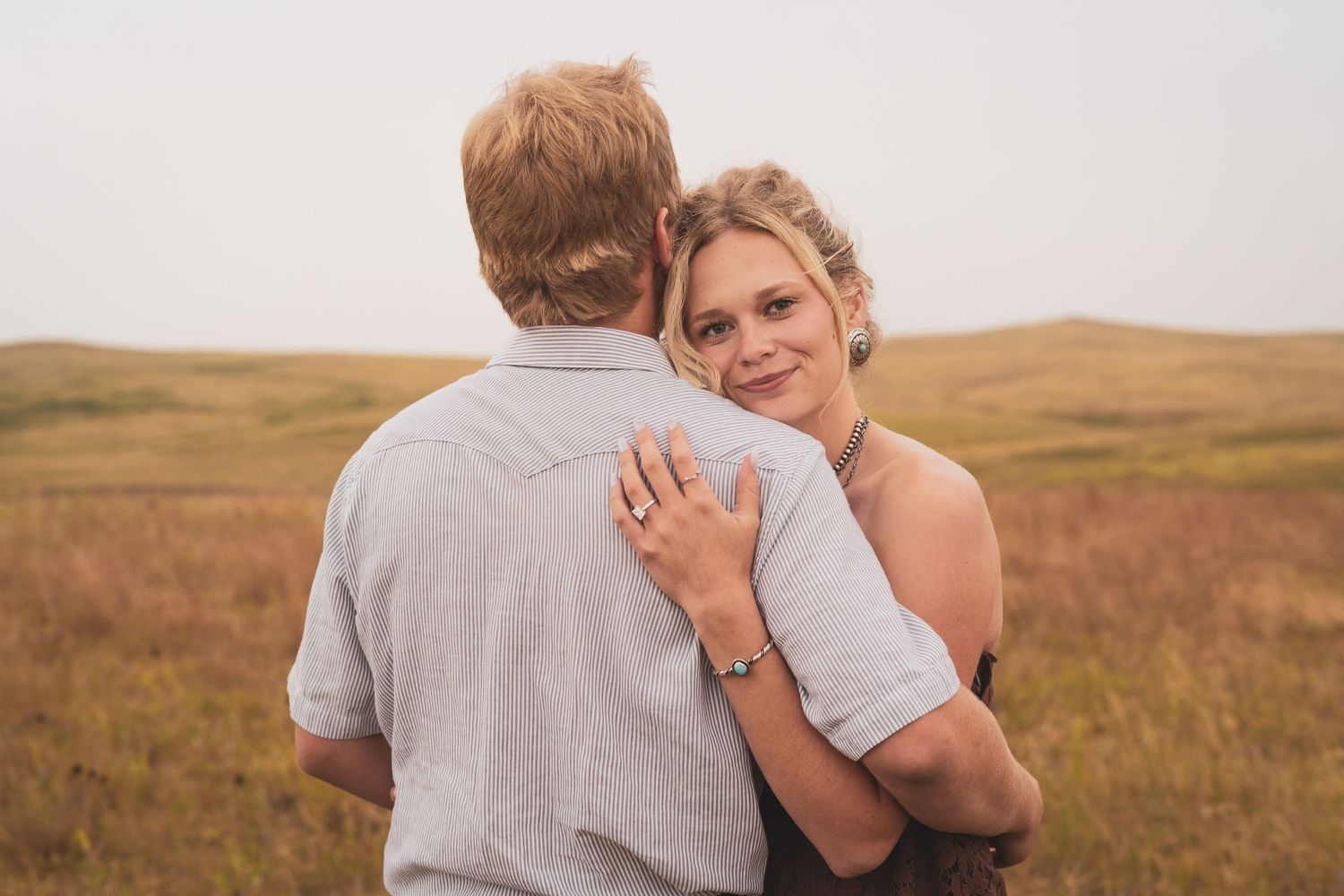 Smiling blonde woman hugging a man from behind in a golden field at sunset wearing a teal bracelet.