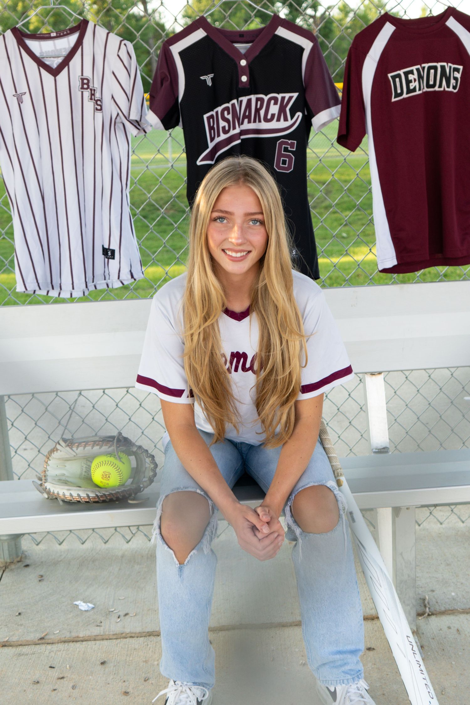 Blonde softball player sitting in dugout with Bismarck Demons jerseys hanging on fence behind her.
