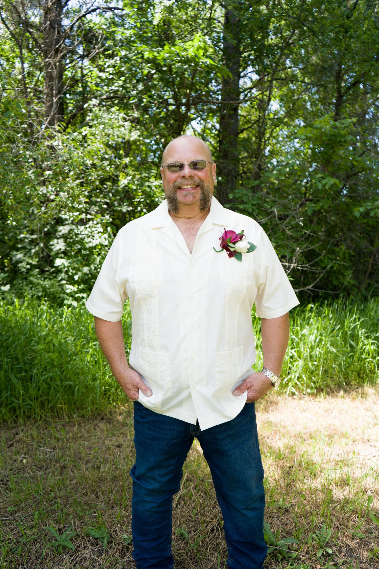 Smiling bald groom in cream short-sleeve shirt with pink boutonniere posing outdoors in lush green setting.