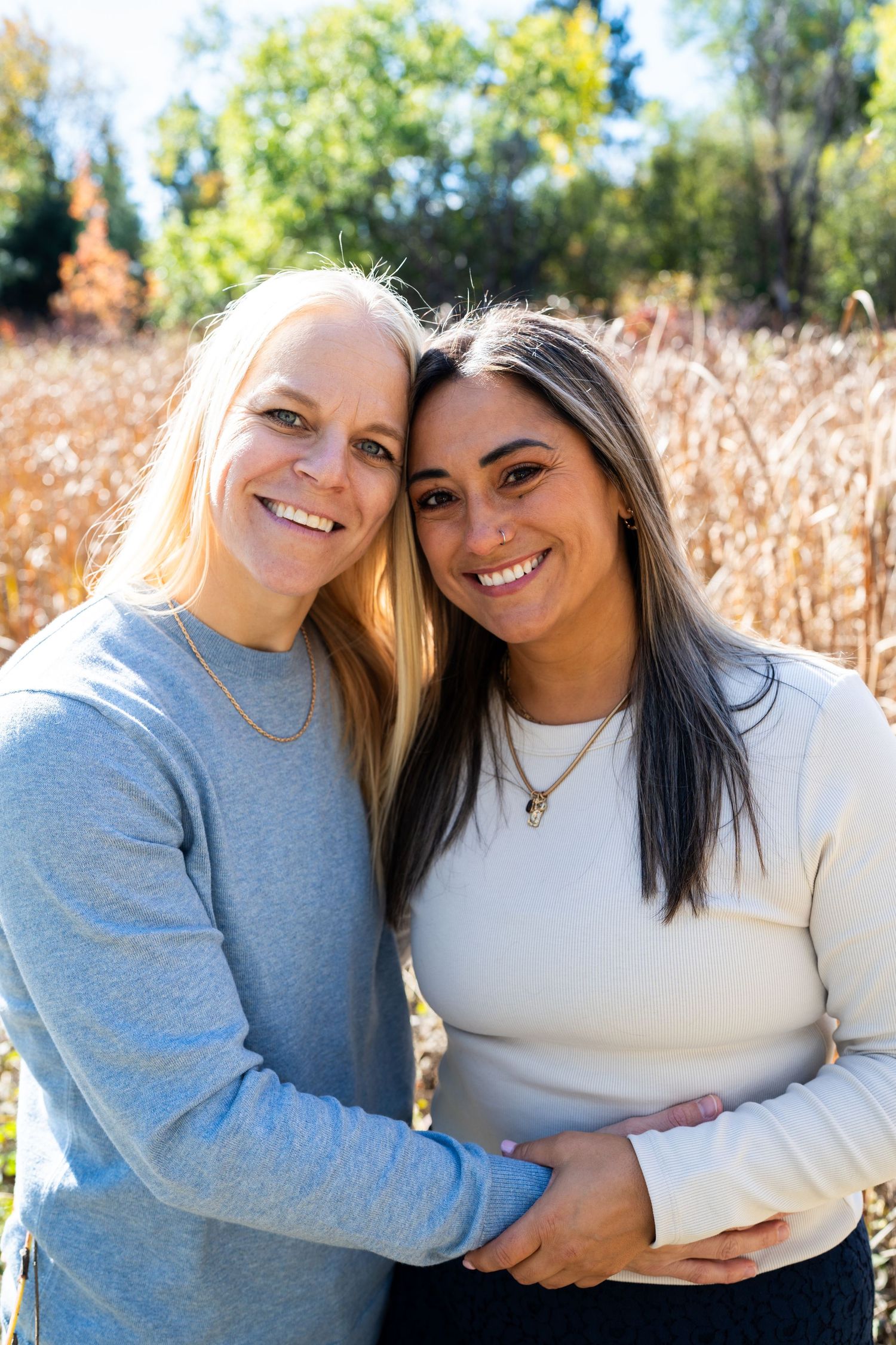 Two smiling women embrace outdoors in front of autumn grasses.