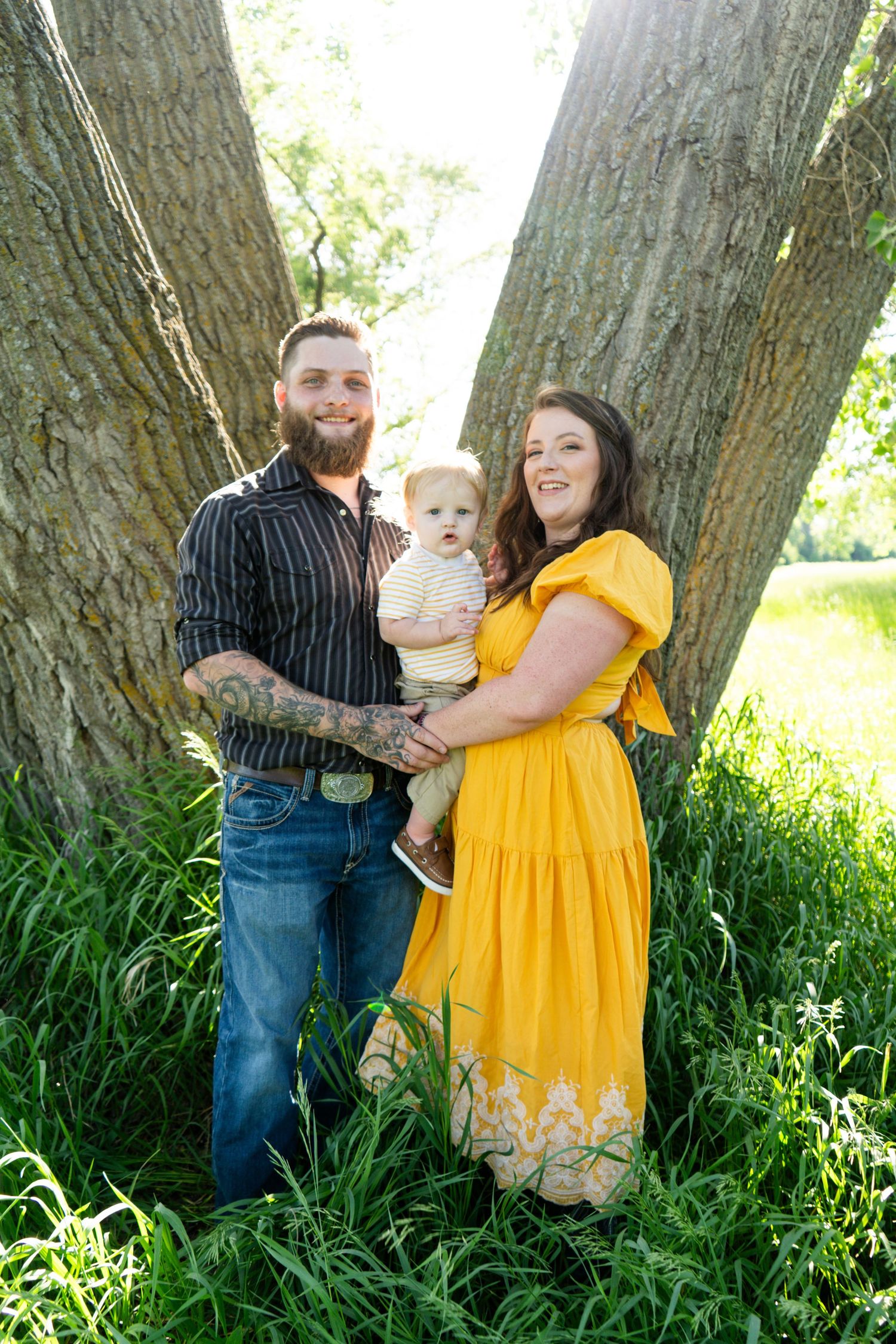A young family with a tattooed father, mother in yellow dress, and baby pose between two trees.