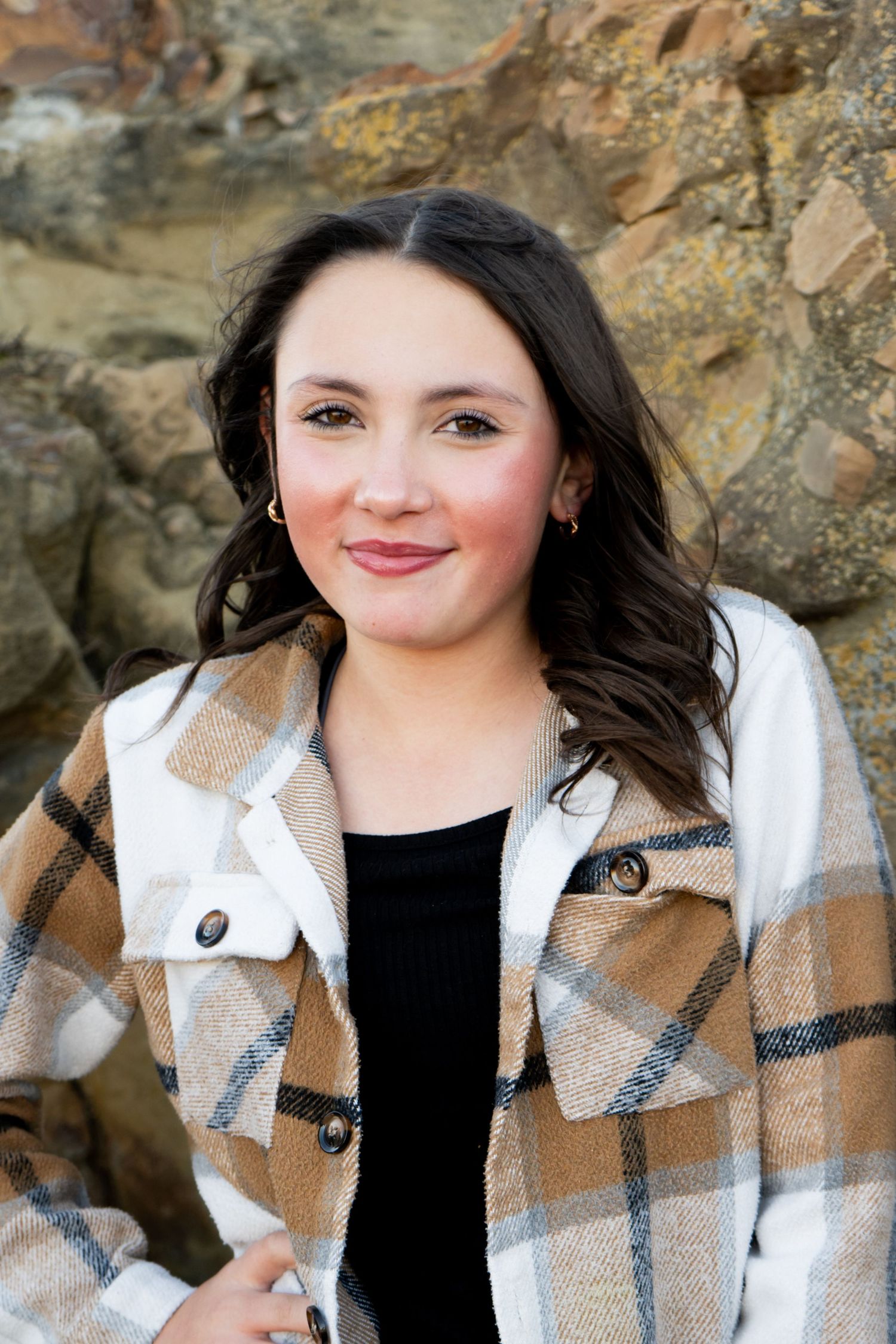 Teenage girl with dark wavy hair smiling in plaid flannel jacket against stone wall.