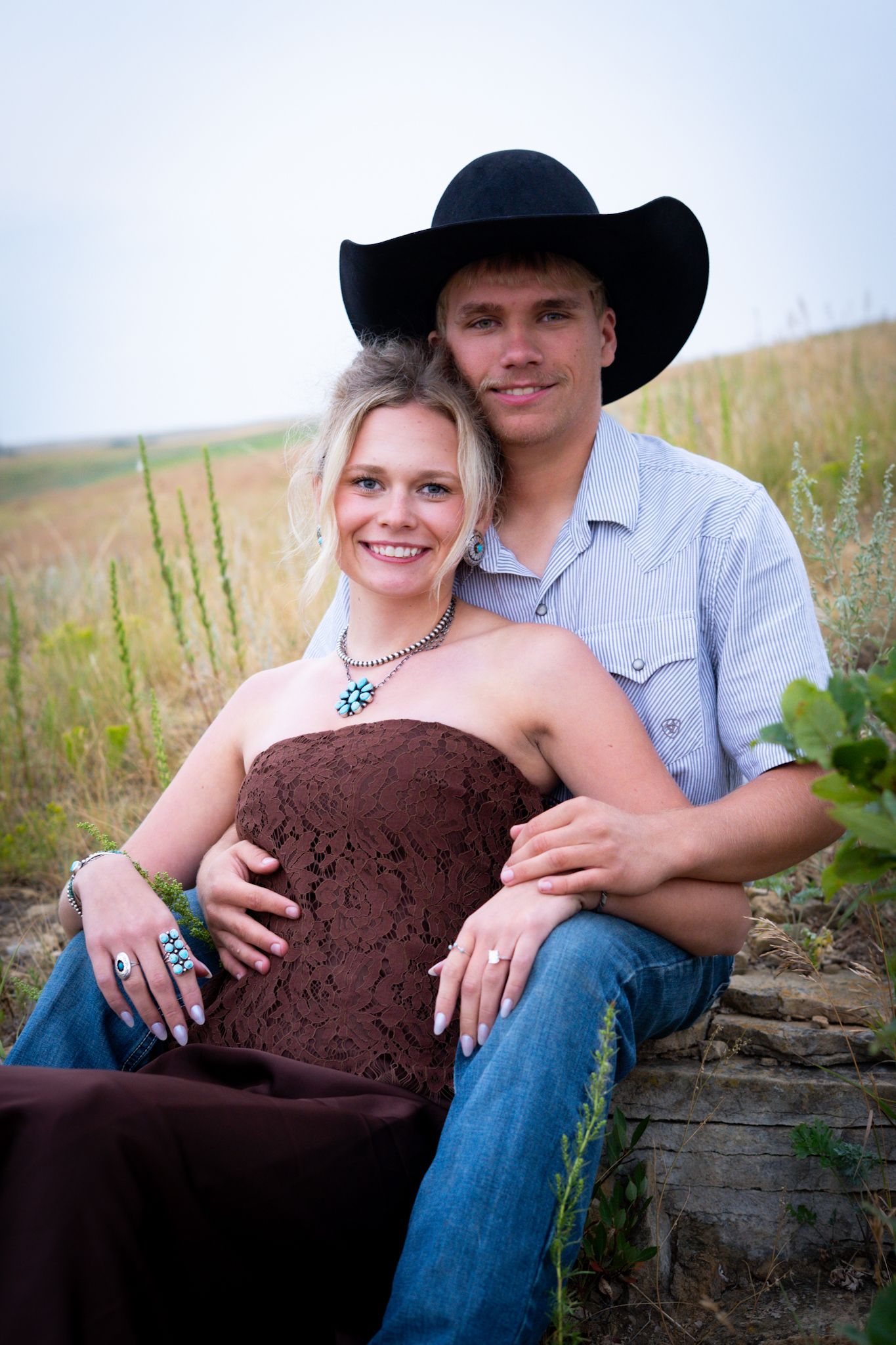 Smiling couple sitting together in a field, man in black cowboy hat embracing blonde woman in brown strapless dress.