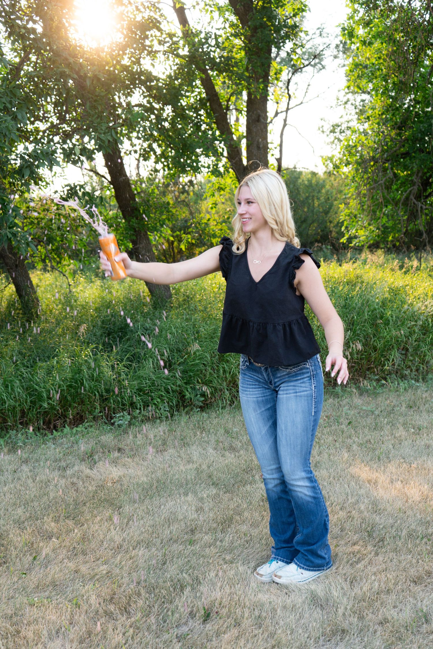 Blonde woman in black top and jeans sprays water from bottle outdoors at sunset.