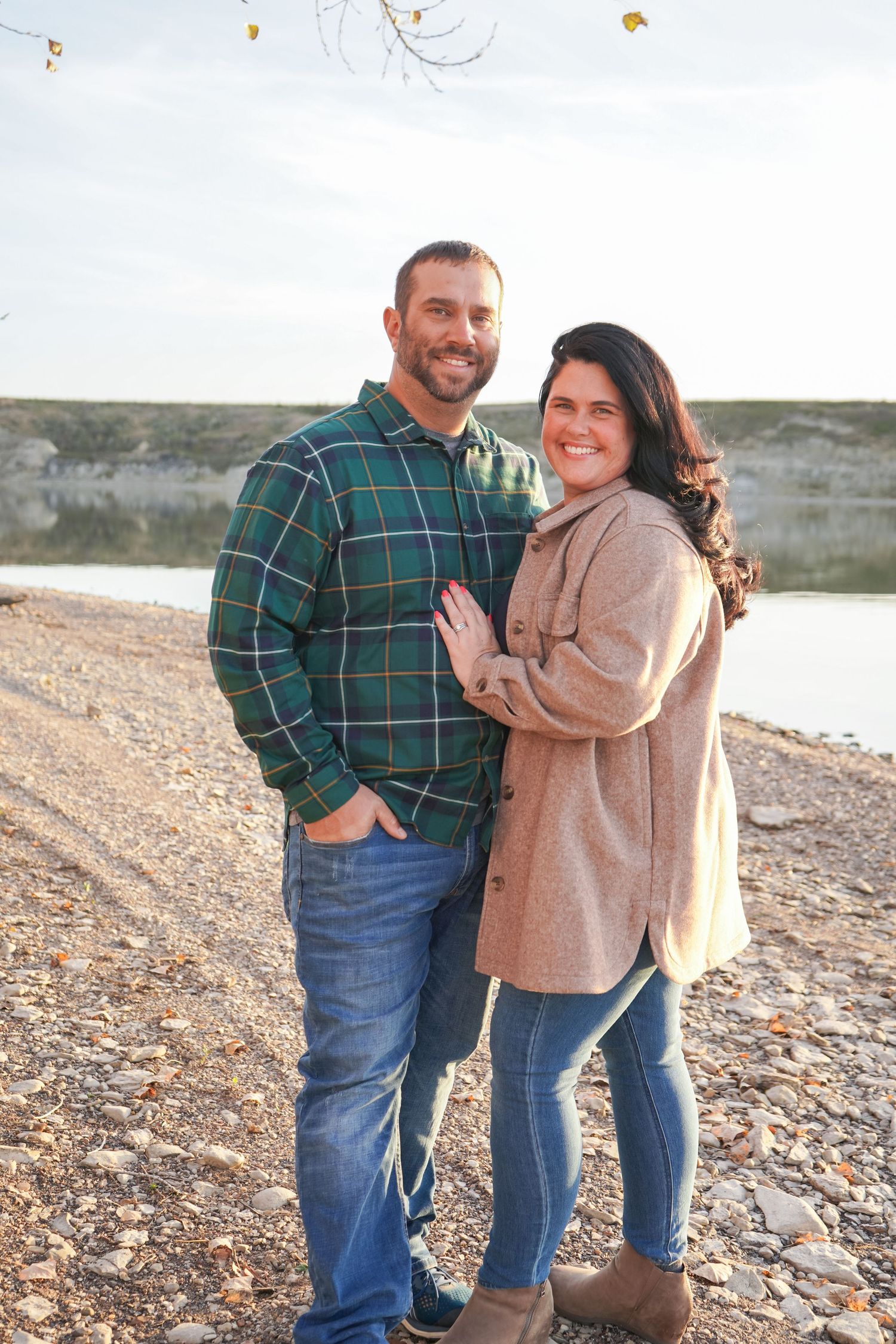 Smiling couple posing by a rocky shoreline in autumn, man in plaid shirt and woman in tan coat.