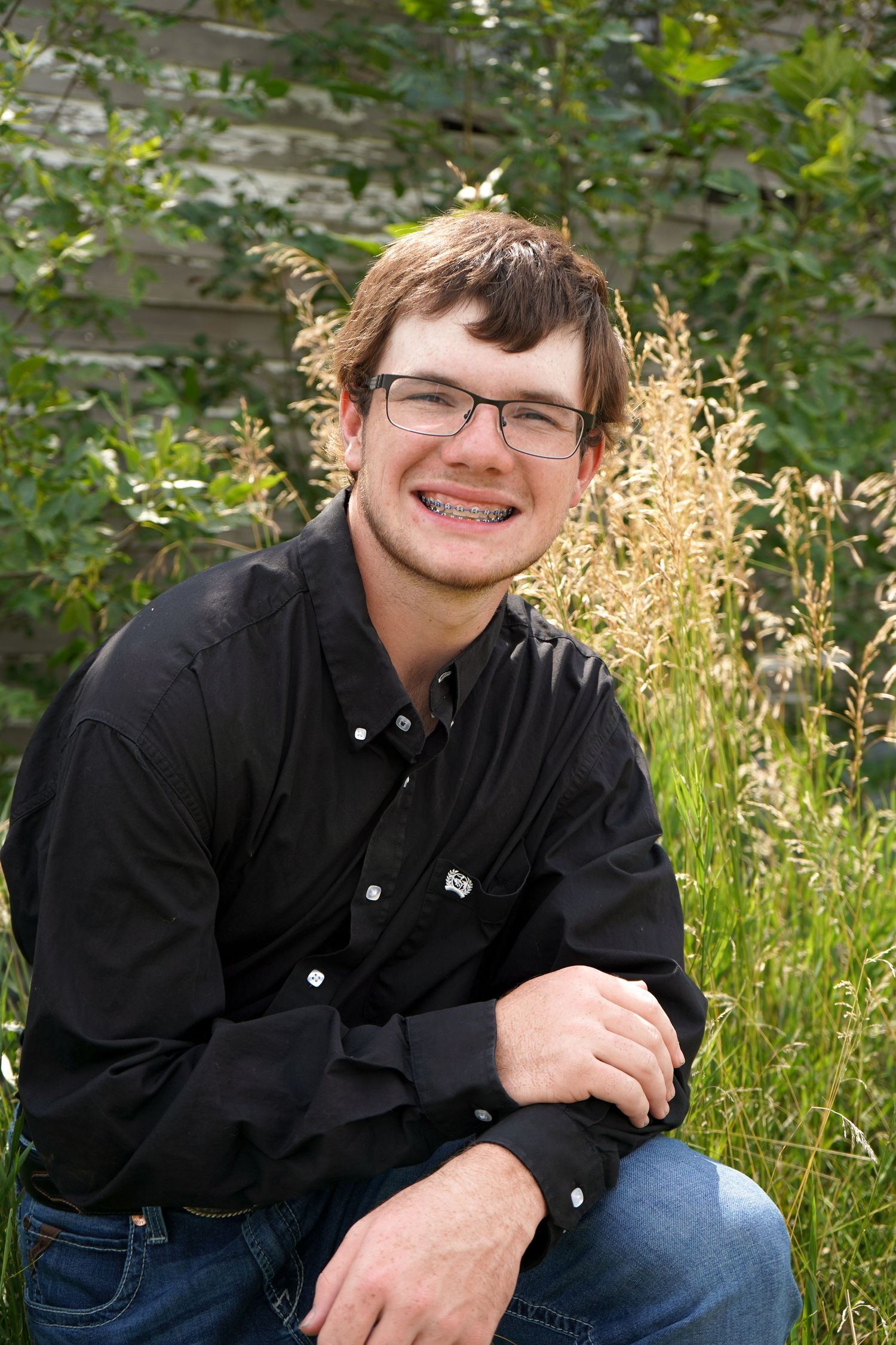 Smiling teen boy with braces and glasses kneeling in green grass near rustic wall.