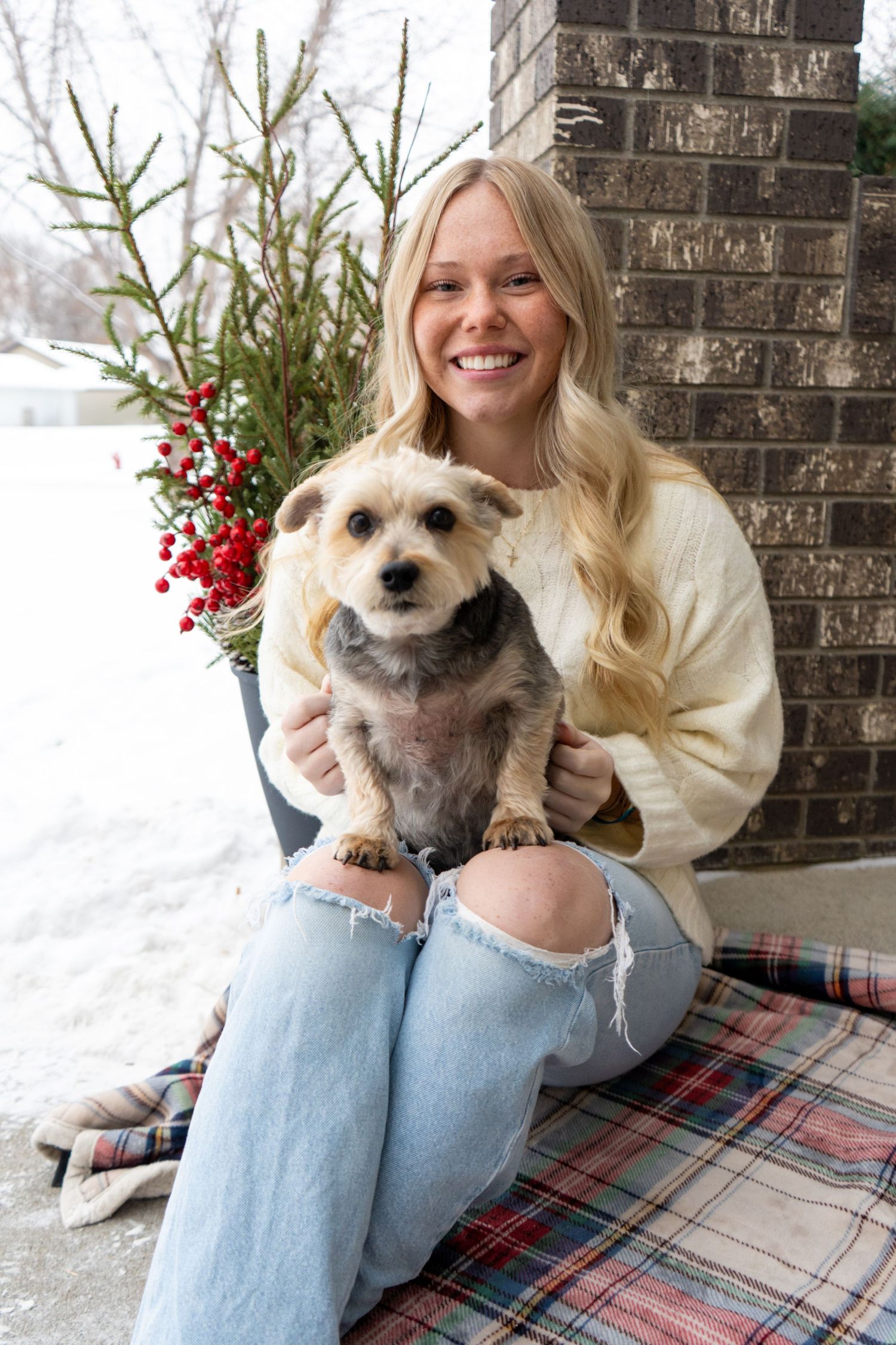 Smiling blonde woman sitting outdoors in winter holding small terrier dog on plaid blanket.